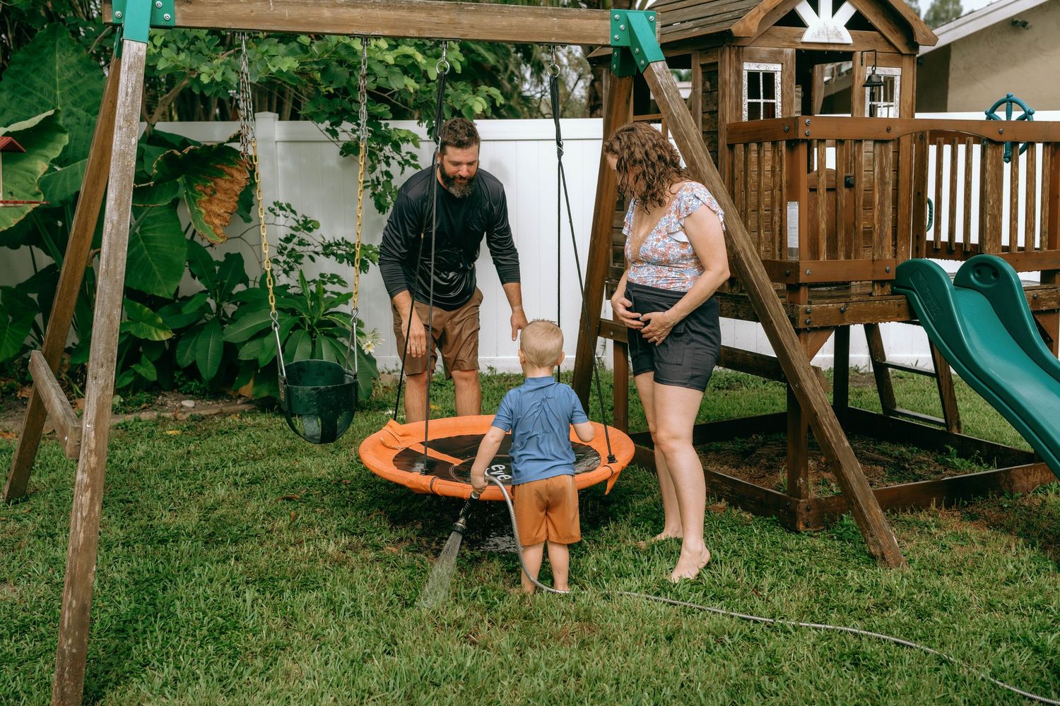 Family playing with toddler on orange saucer swing in backyard with wooden playset and green slide.