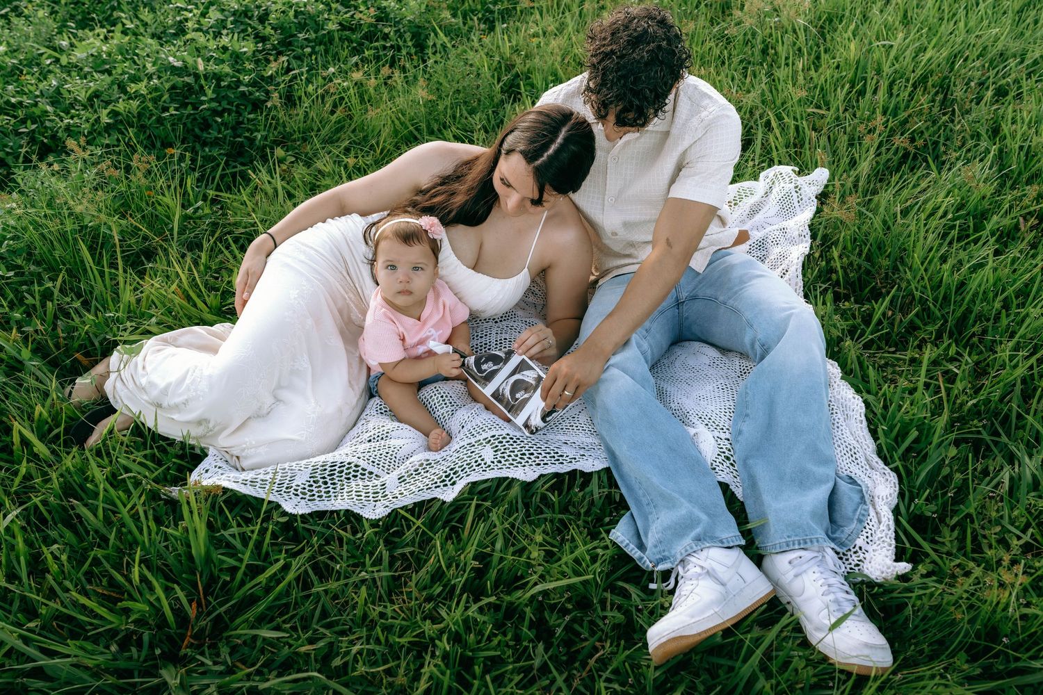 Family of three sitting on blanket in green grass, parents looking at ultrasound photo with baby.