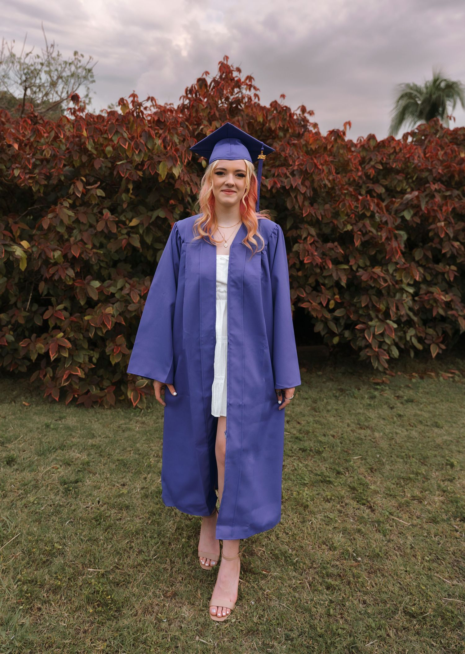 A graduate poses in traditional blue cap and gown against autumn foliage.