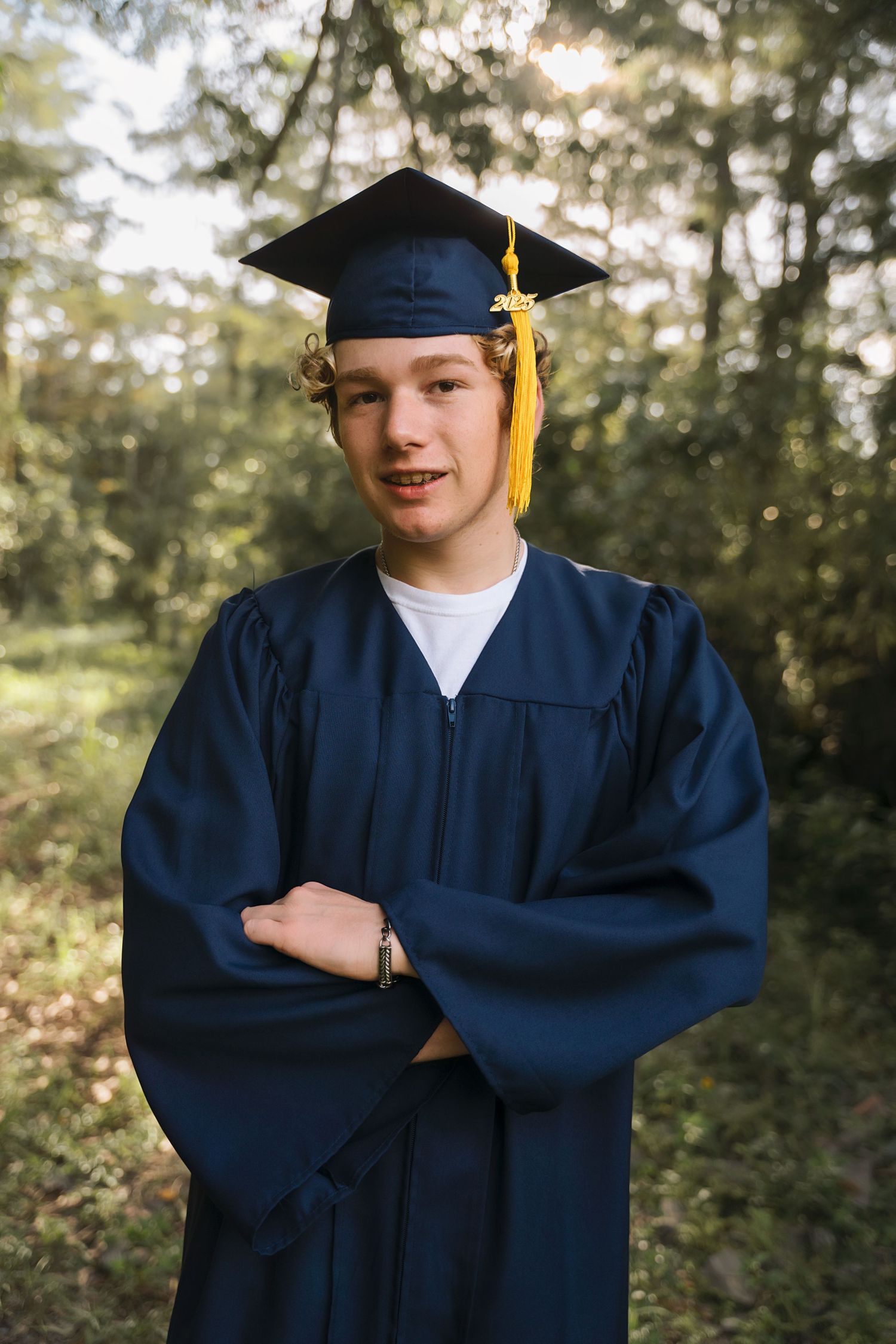 A graduate in navy blue cap and gown poses in a forest setting.