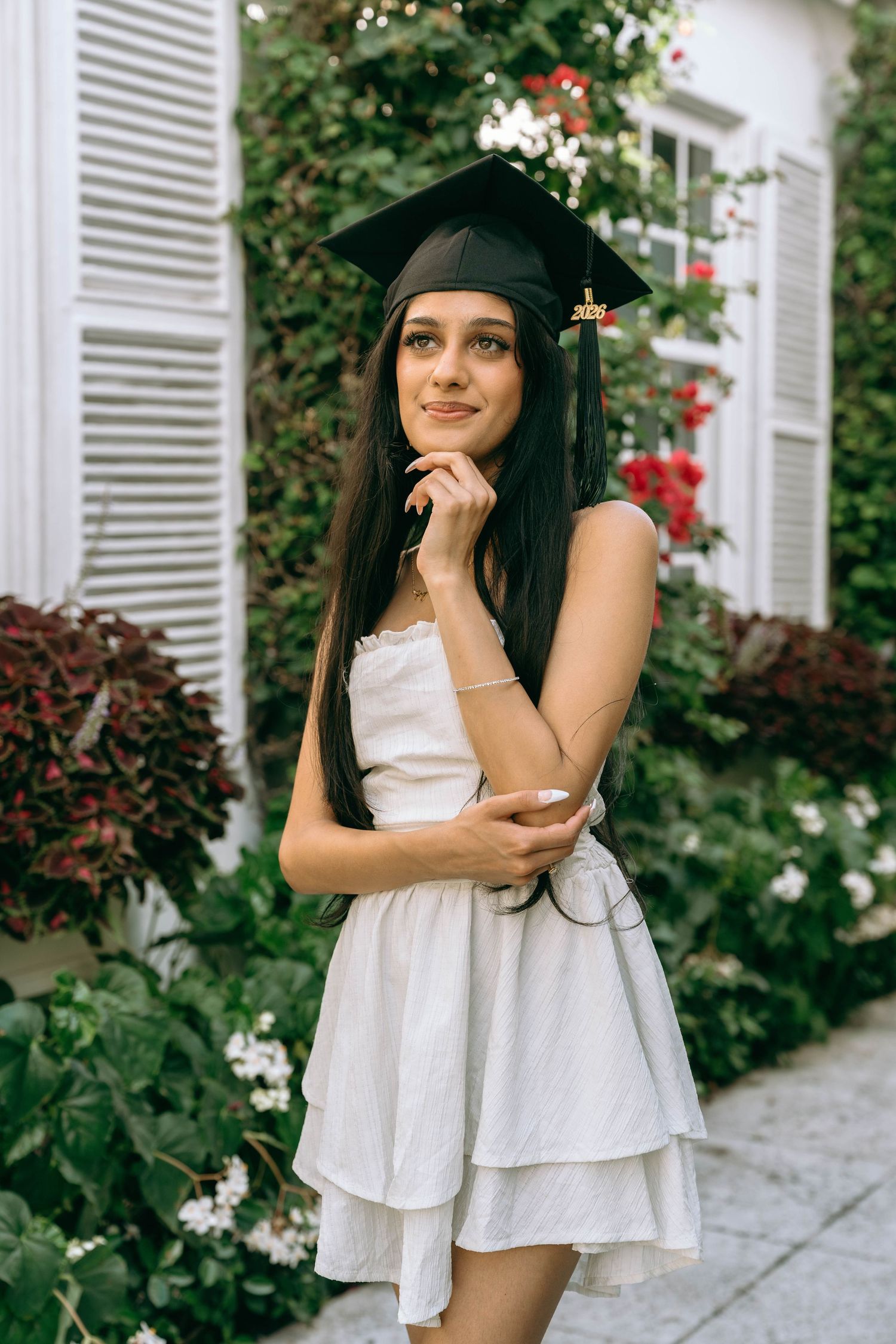 Young woman in white dress and graduation cap posing thoughtfully in a flower garden on Worth Avenue Palm beach.