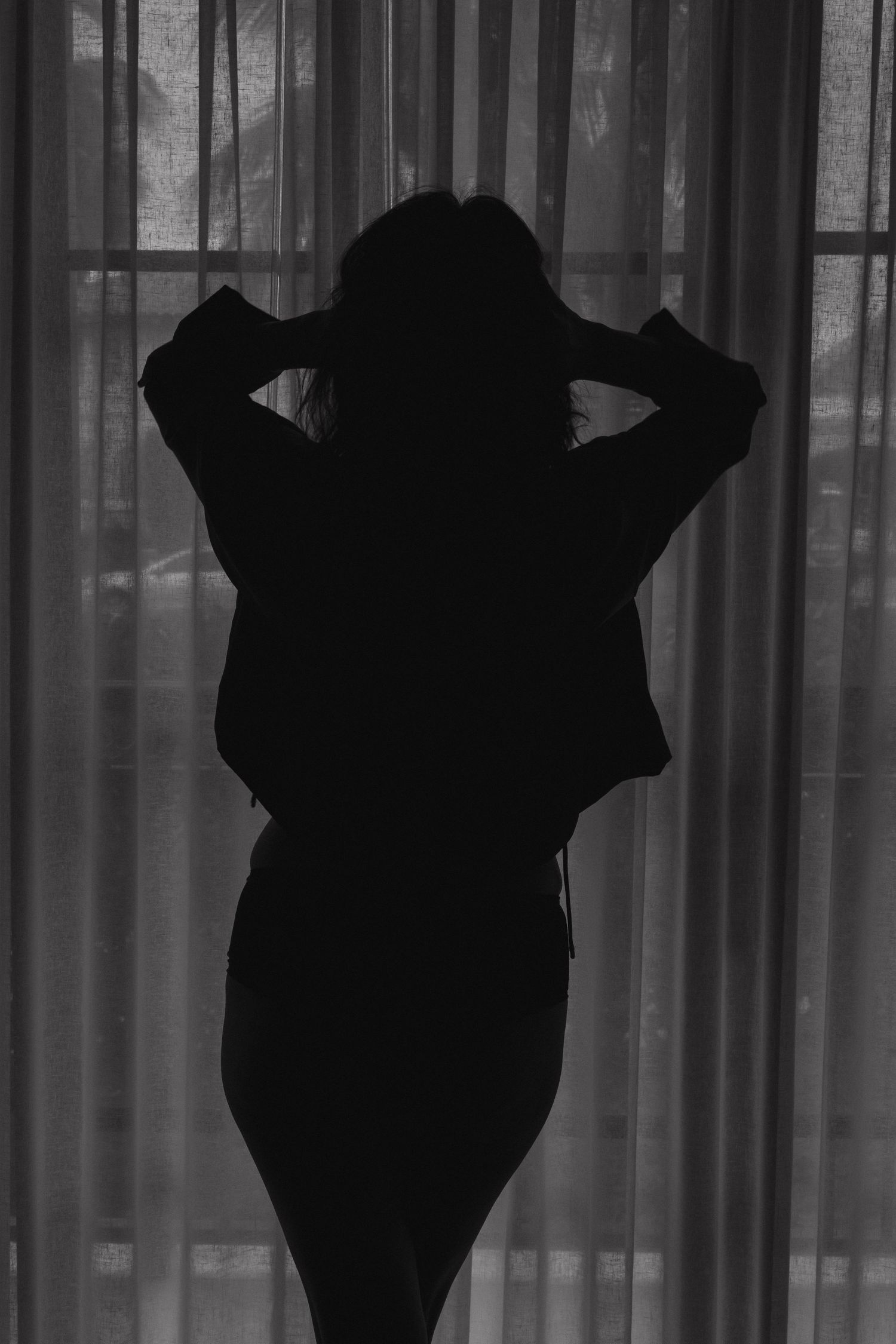 Black and white silhouette of a woman standing before sheer curtains with hands in hair.  Boudoir Palm Beach, FL