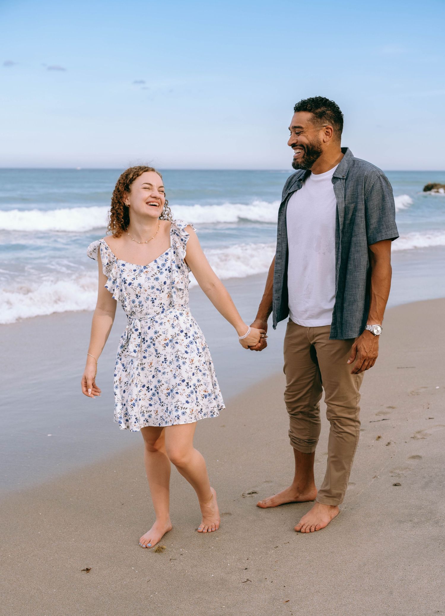 Happy couple holding hands and walking barefoot on a sandy beach with ocean waves in the background. Engagement photos on Worth Avenue