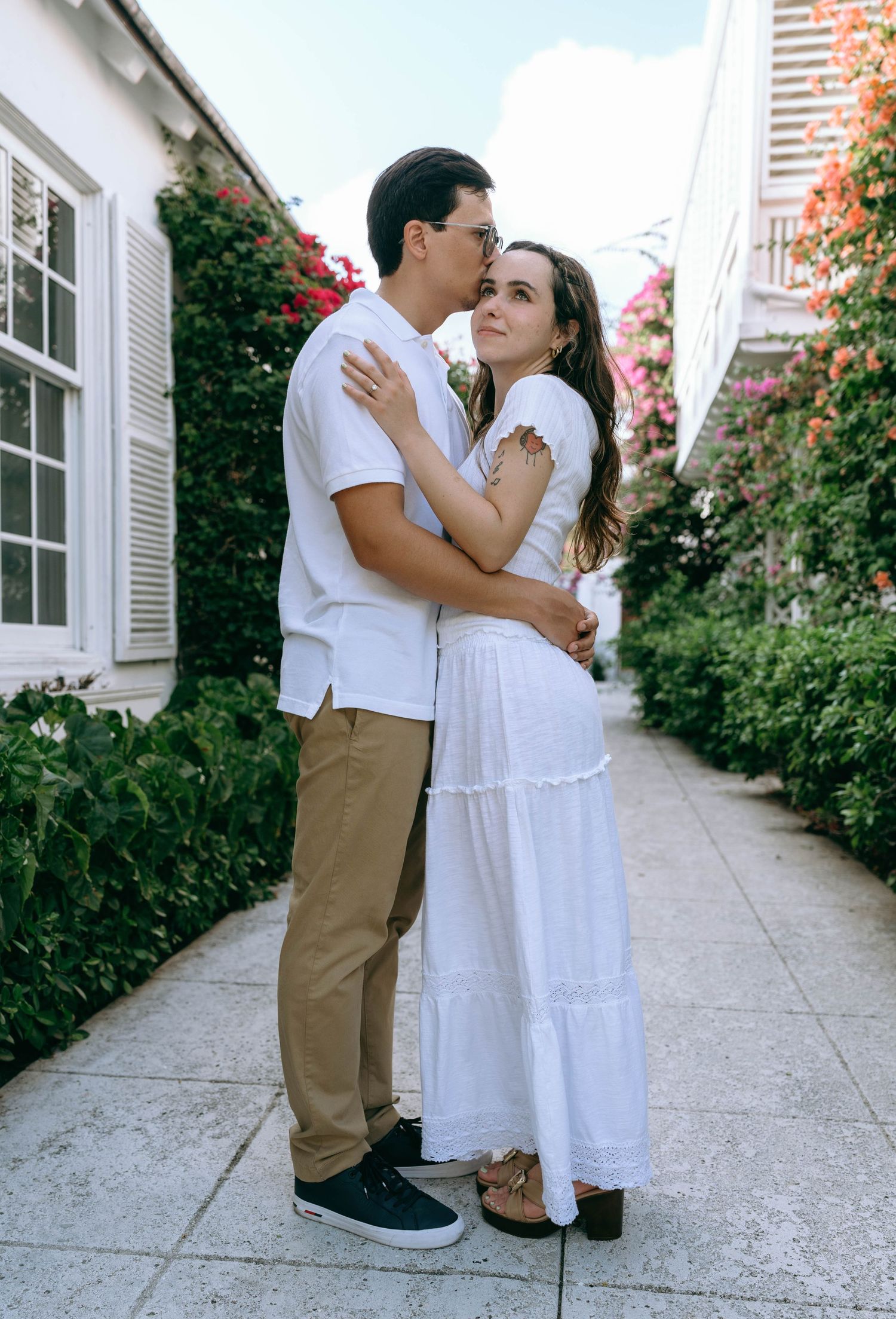 A couple in matching white outfits pose romantically in a sunlit garden corridor with pink flowers. Engagement photos on Worth Avenue
