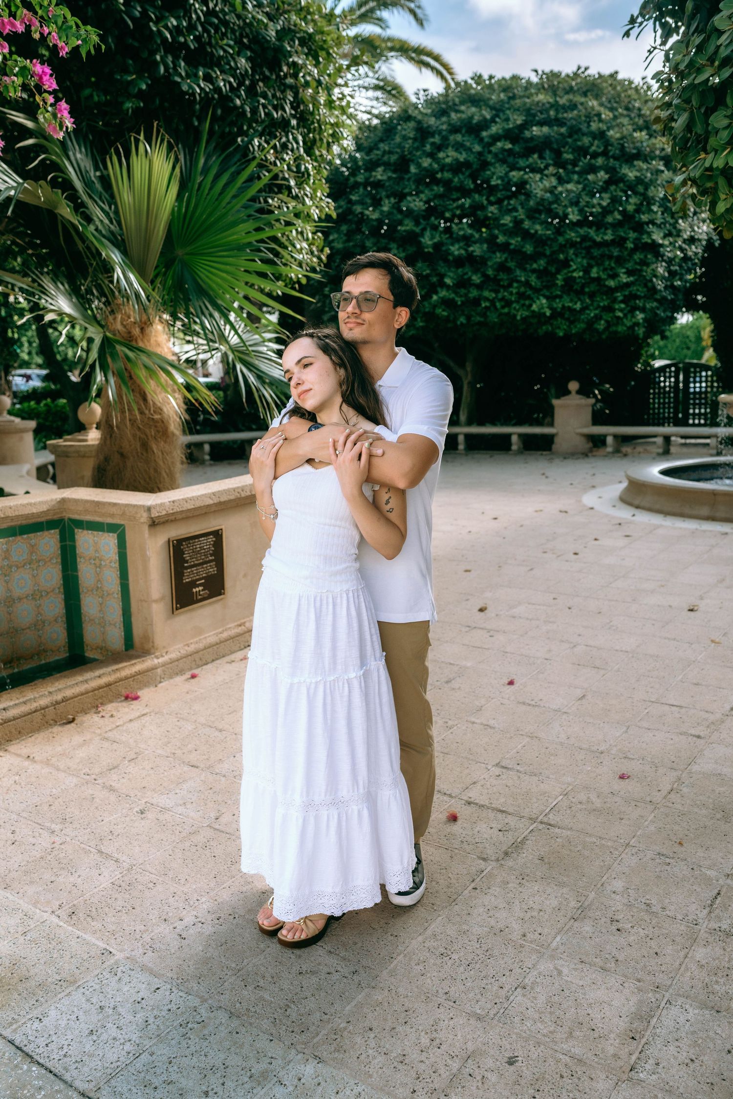 A couple embracing in a sunny garden, woman in white dress, man in white shirt and khakis, surrounded by lush greenery. Worth Avenue,