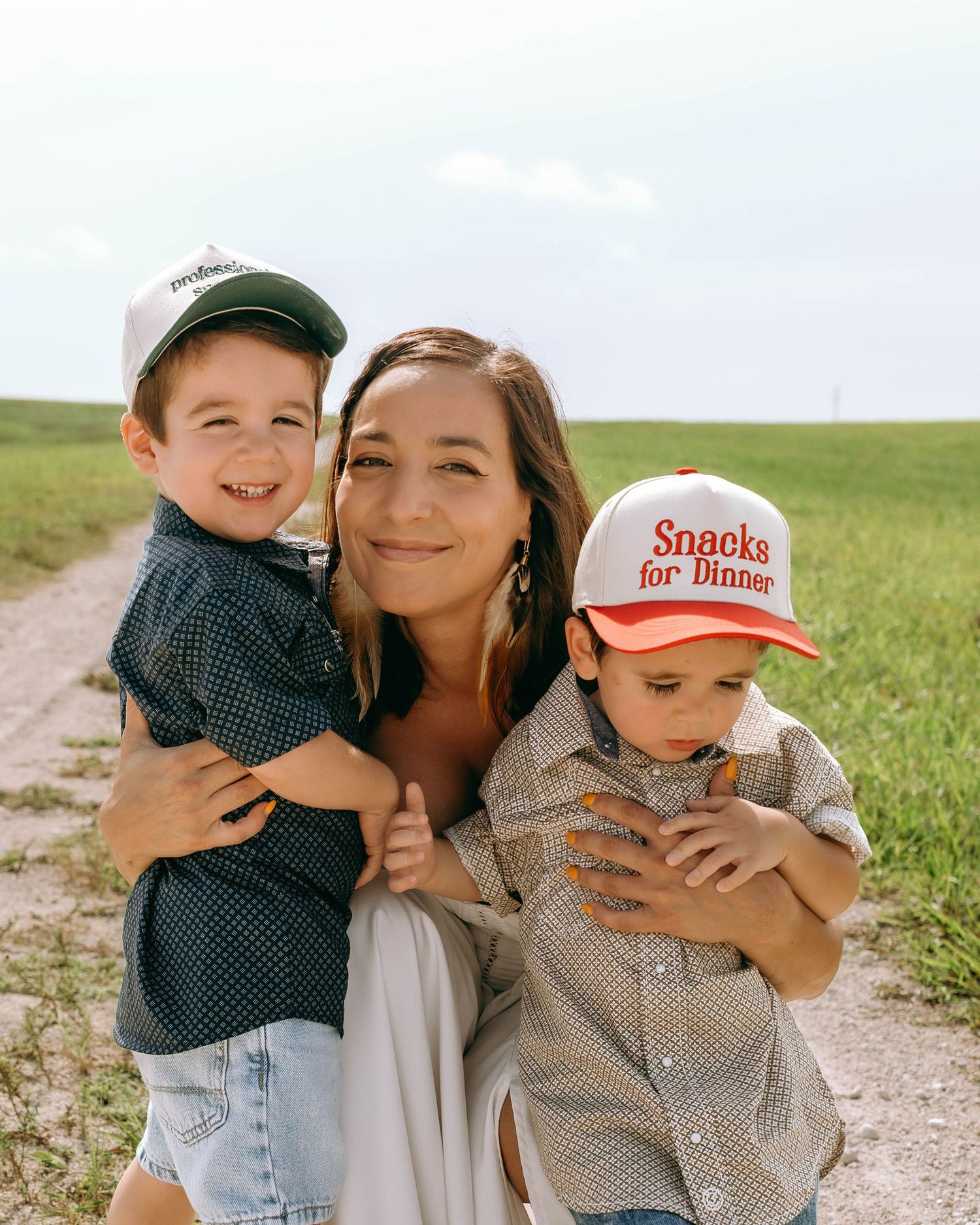 A woman smiles warmly while hugging two young boys on a rural dirt path surrounded by green fields.