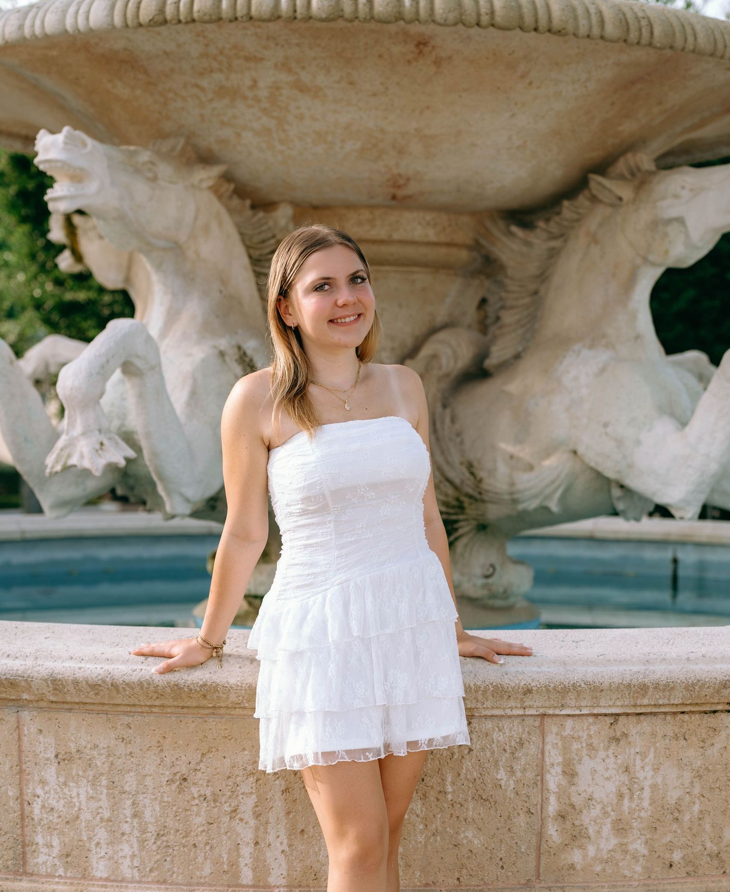 Young woman in white strapless mini dress posing by ornate stone fountain with horse sculptures.