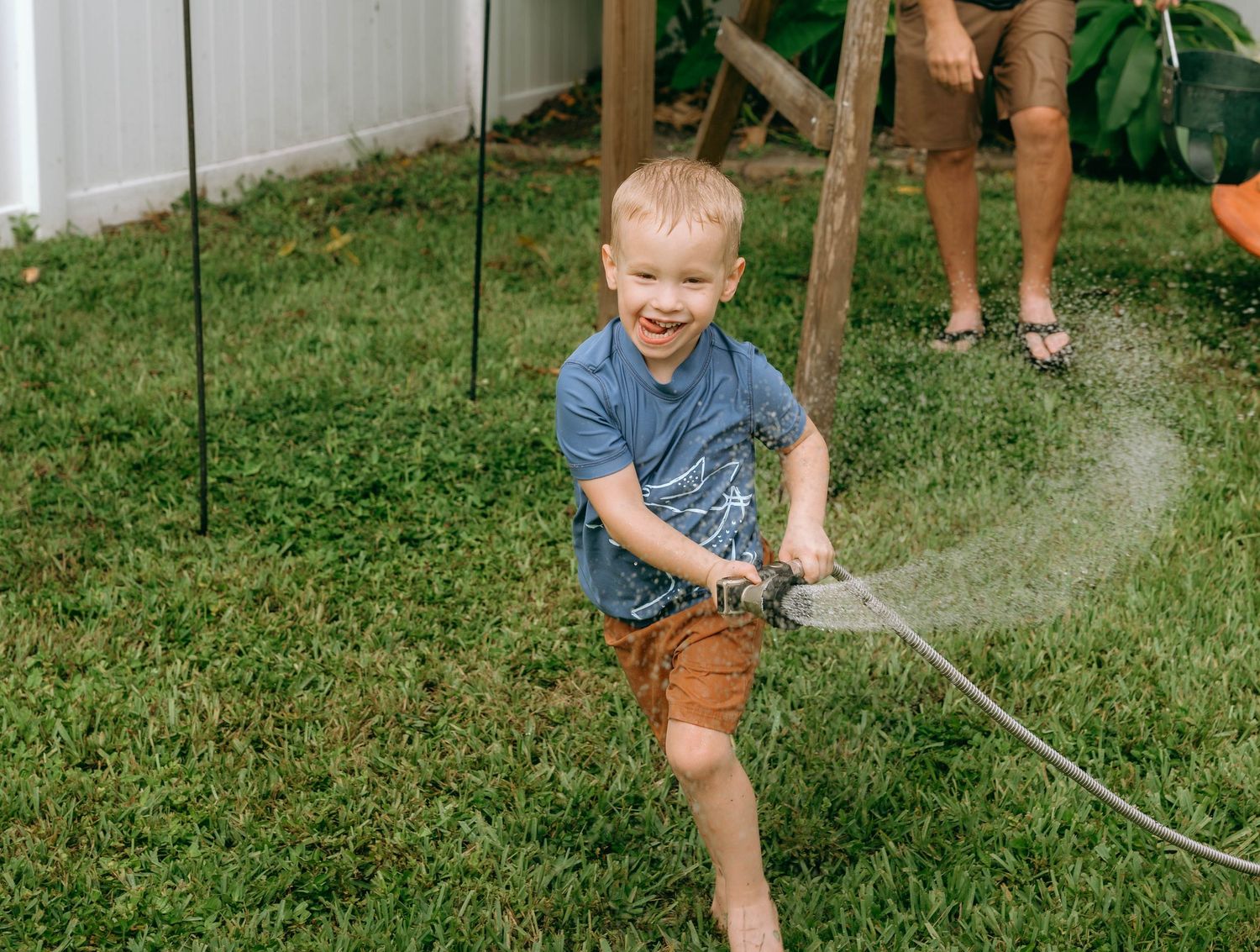 Young boy laughing while spraying water with garden hose on green lawn during summer outdoor play.