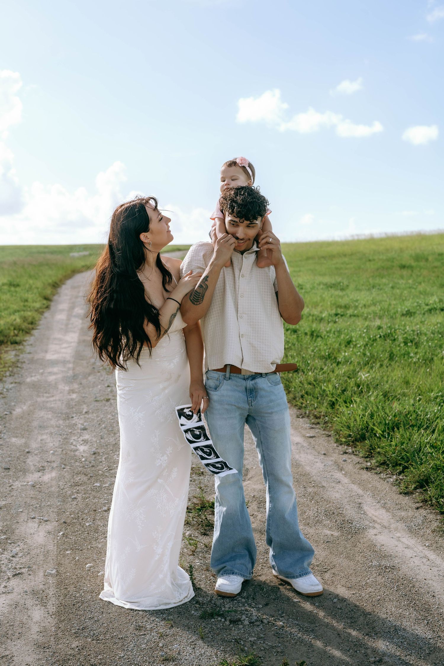 A happy family poses on a rural dirt path, with mom in white dress, dad in jeans holding their young daughter on his shoulders.