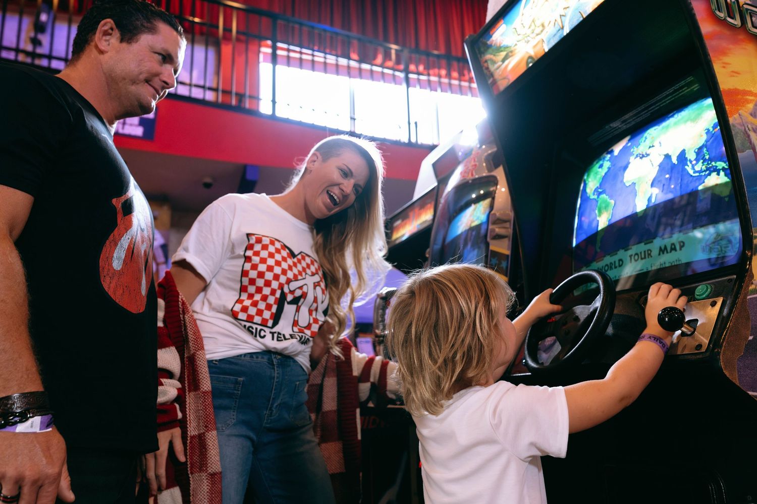 Family enjoying arcade games together, with a child steering a racing game while parents watch and smile.