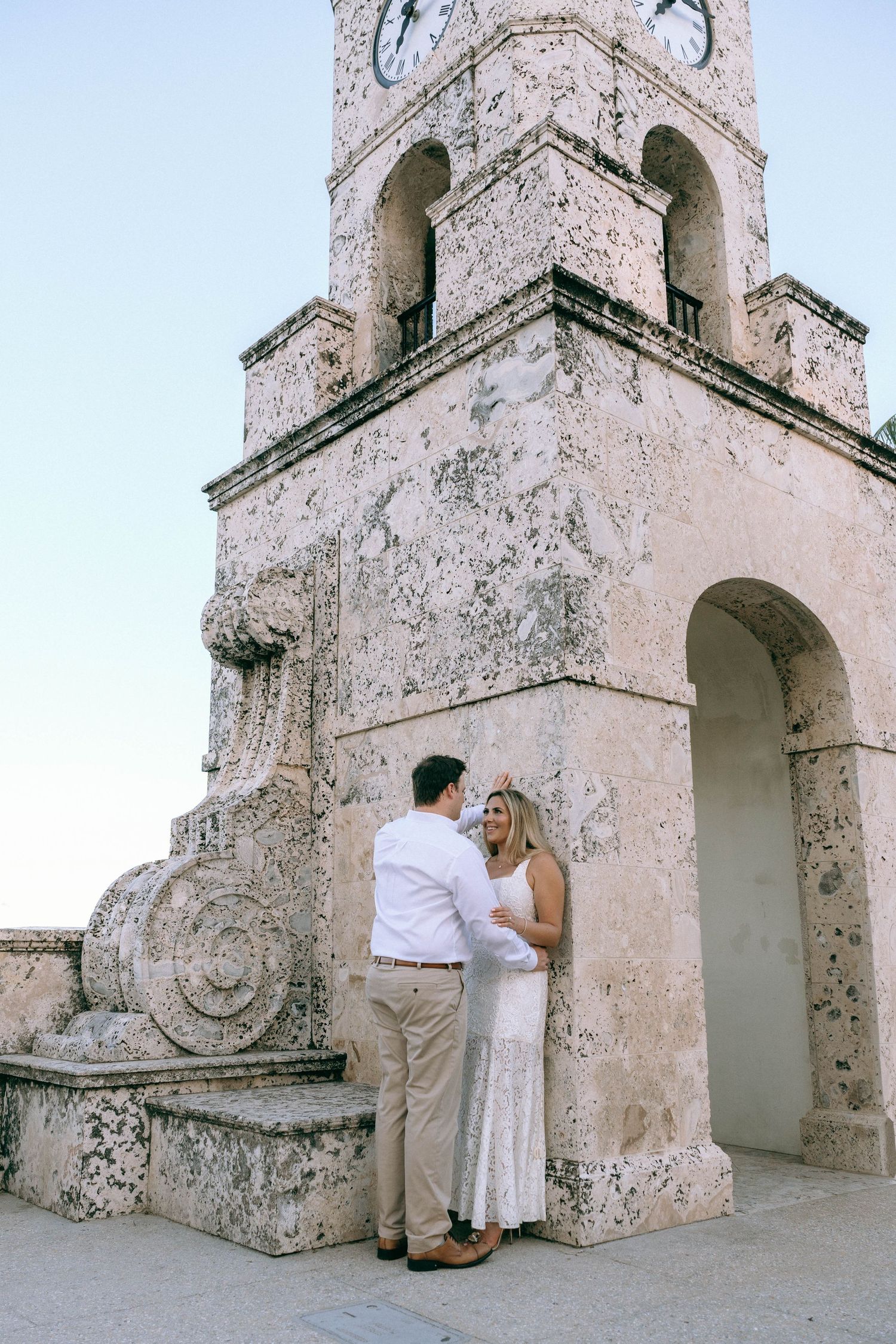 Couple holding hands and gazing at each other beside a historic stone clock tower.