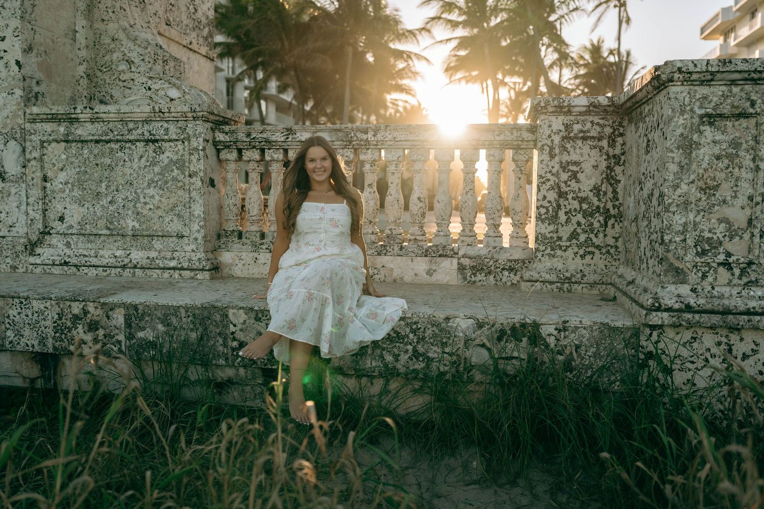 Woman in white sundress standing by stone balustrade with sunset rays through palm trees.