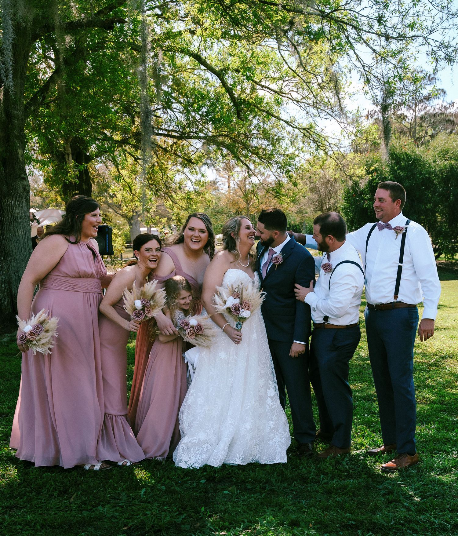 Wedding party poses outdoors under trees, bridesmaids in dusty rose gowns with bride in lace dress and groomsmen in navy suits.