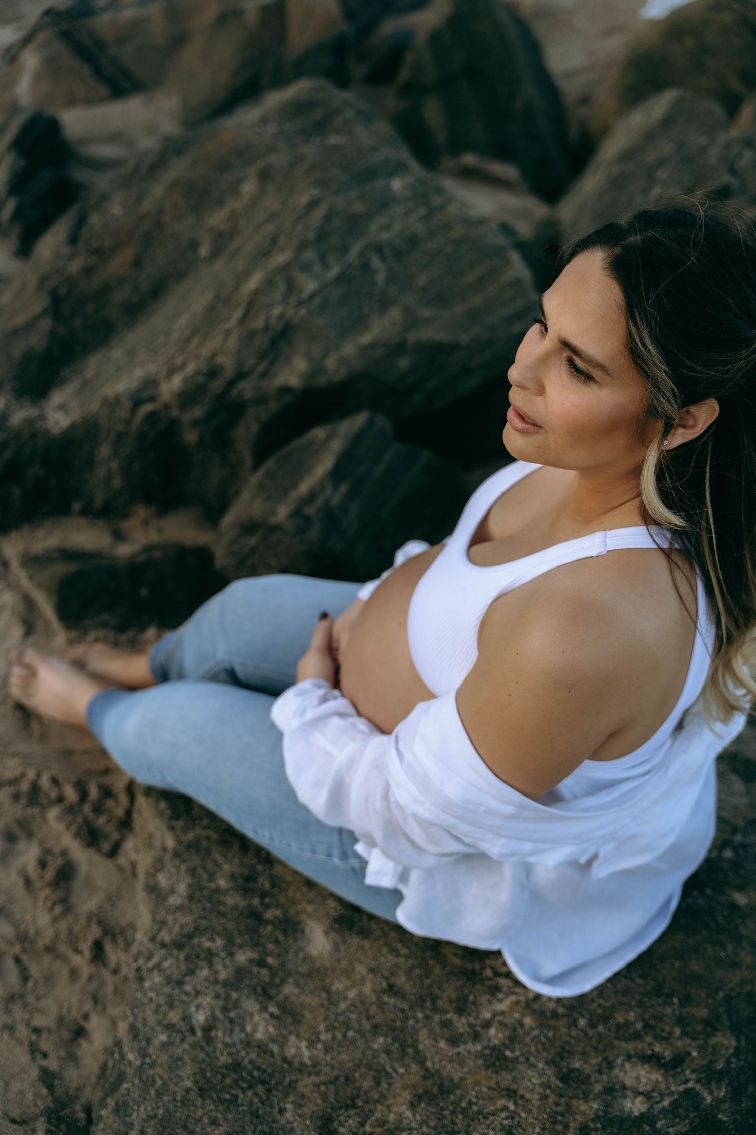 A maternity portrait of someone in white sitting on coastal rocks.