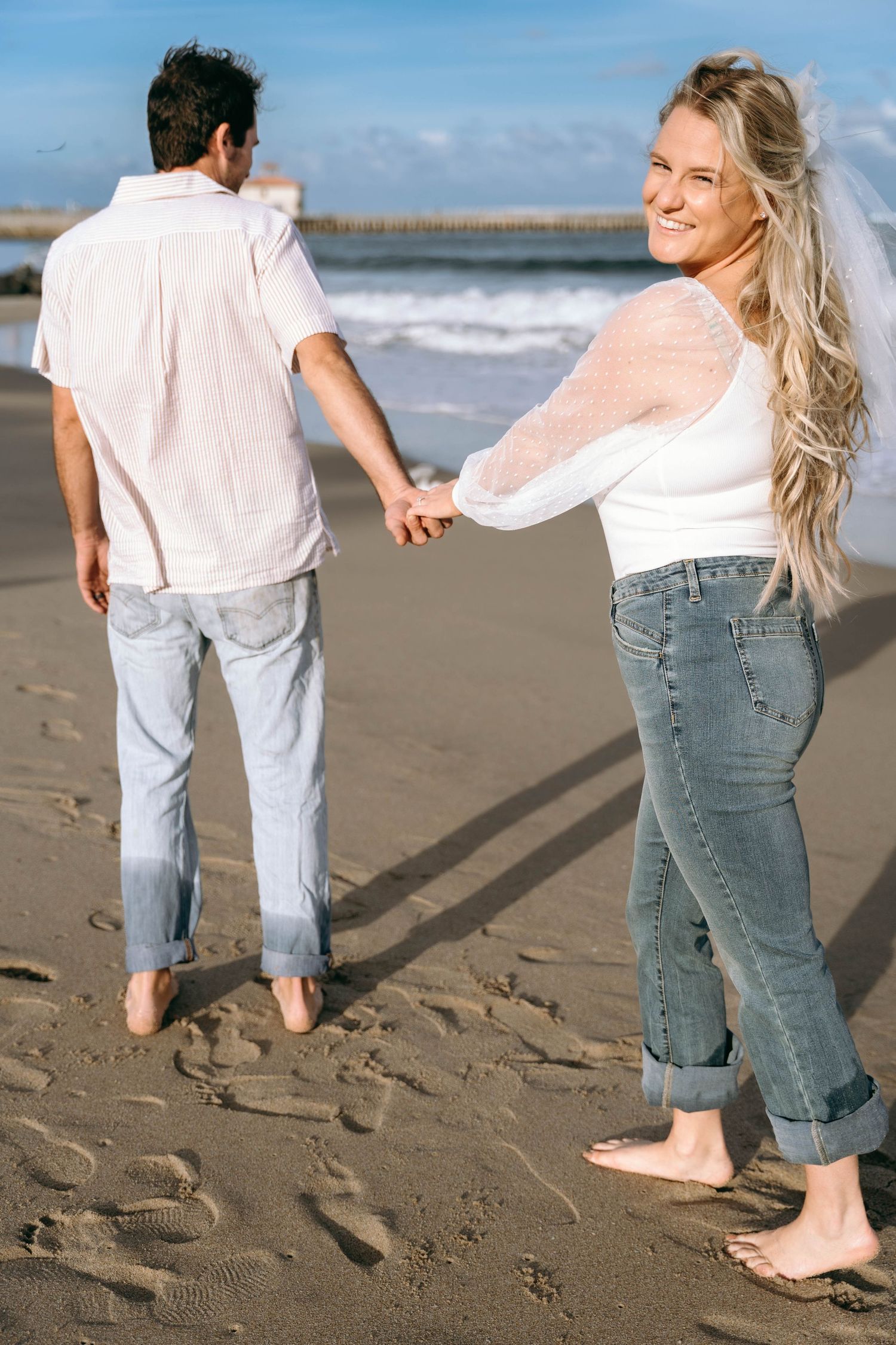 A couple holds hands while walking barefoot on a beach with waves in the background.