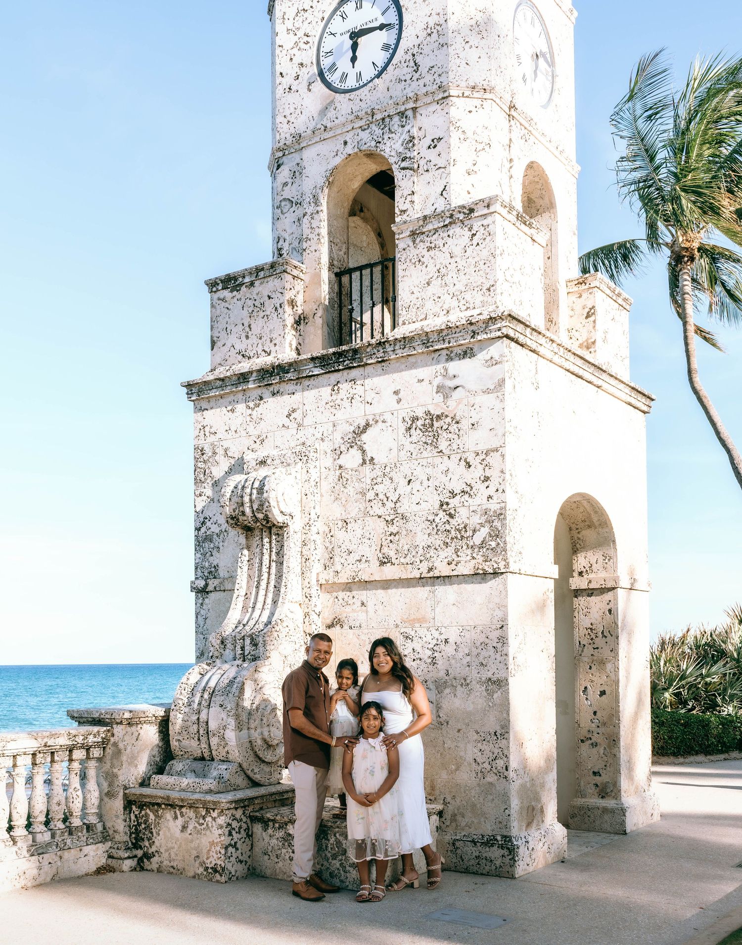 Family of four posing in front of historic Palm Beach clock tower with ocean view and palm trees on Worth Avenue Palm beach.