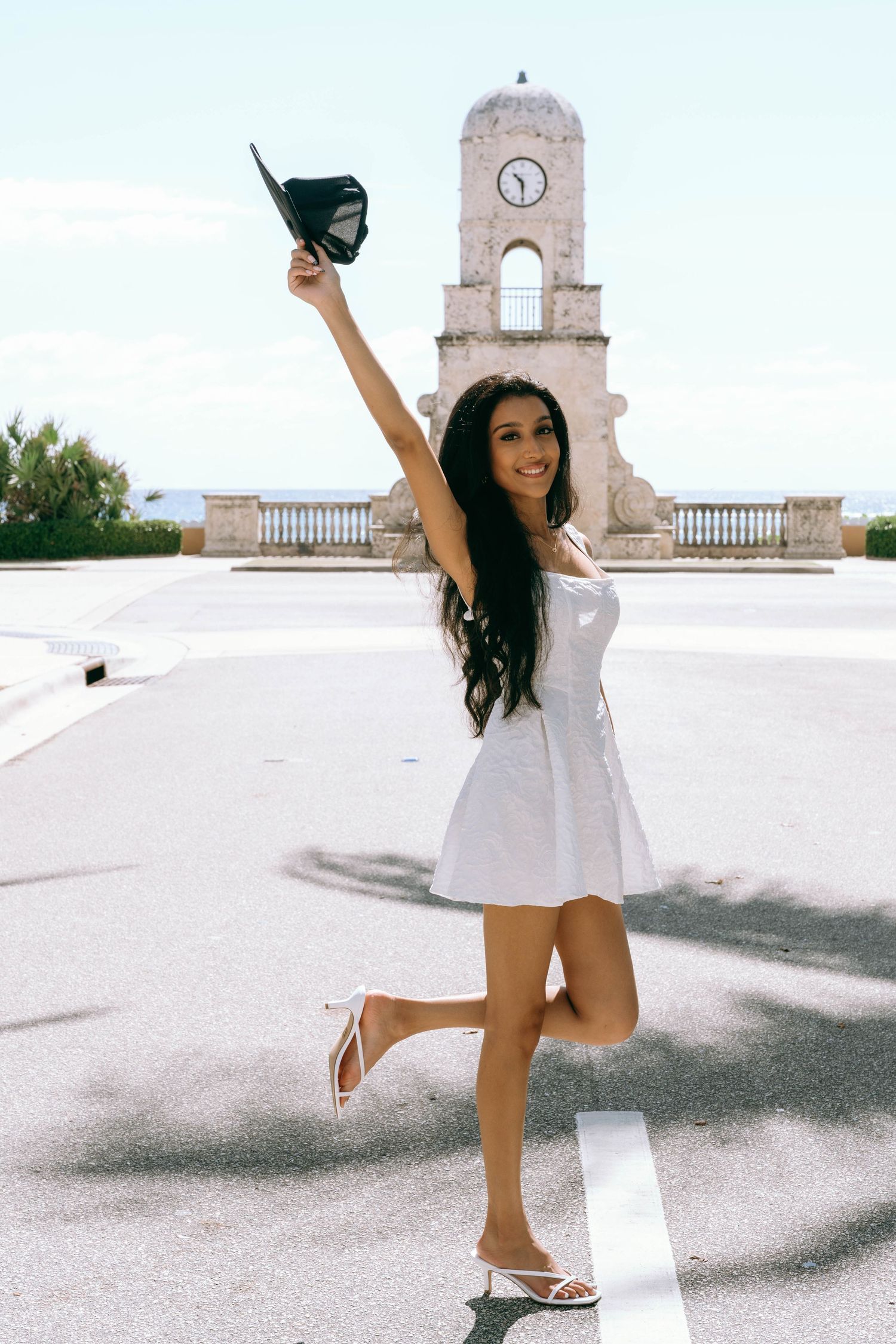 Joyful young woman in white dress raising graduation cap near historic clock tower on Worth Avenue Palm beach.