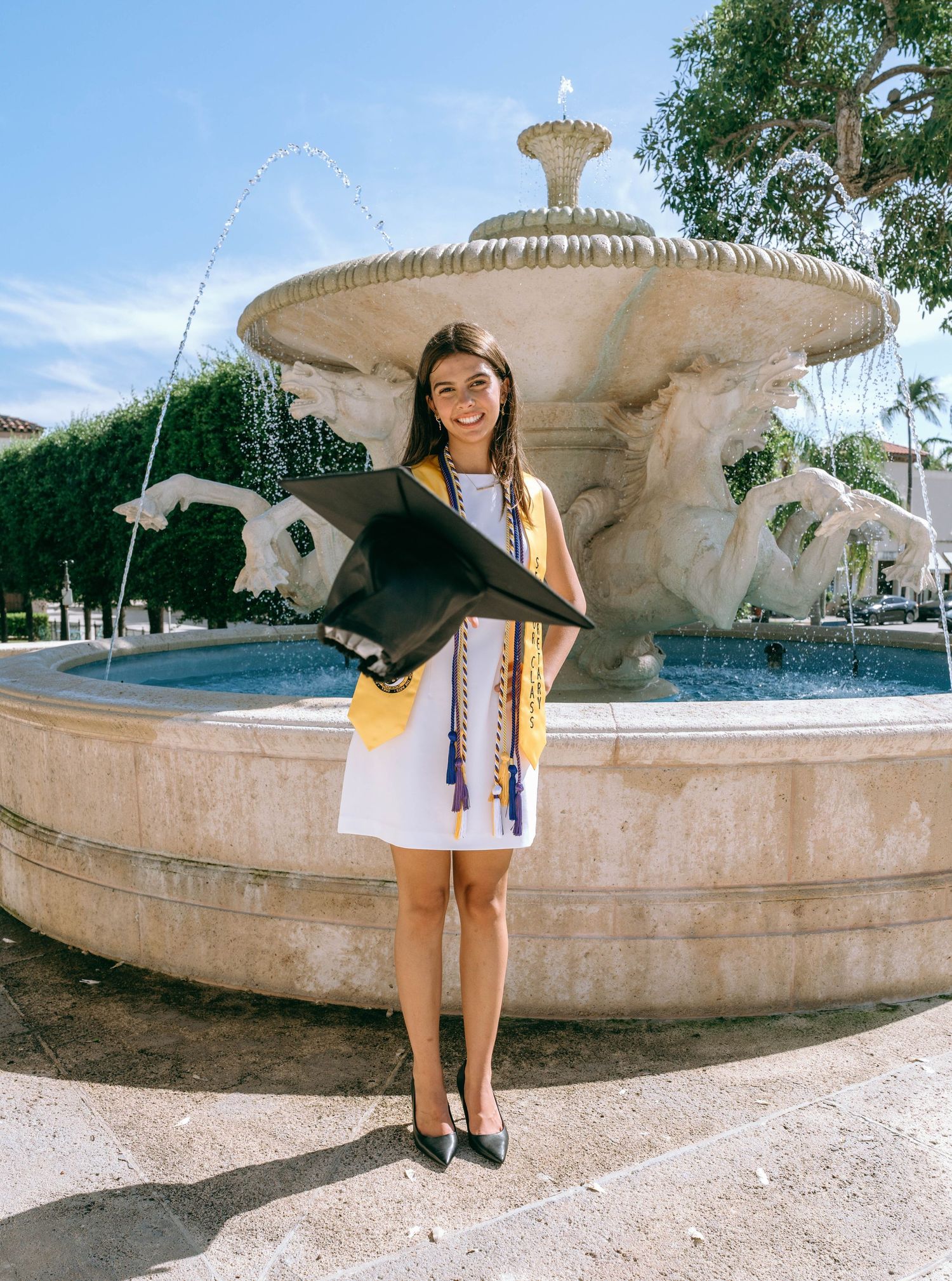 Graduate in cap and gown poses by ornate stone fountain holding diploma on sunny day on Worth Avenue Palm beach
