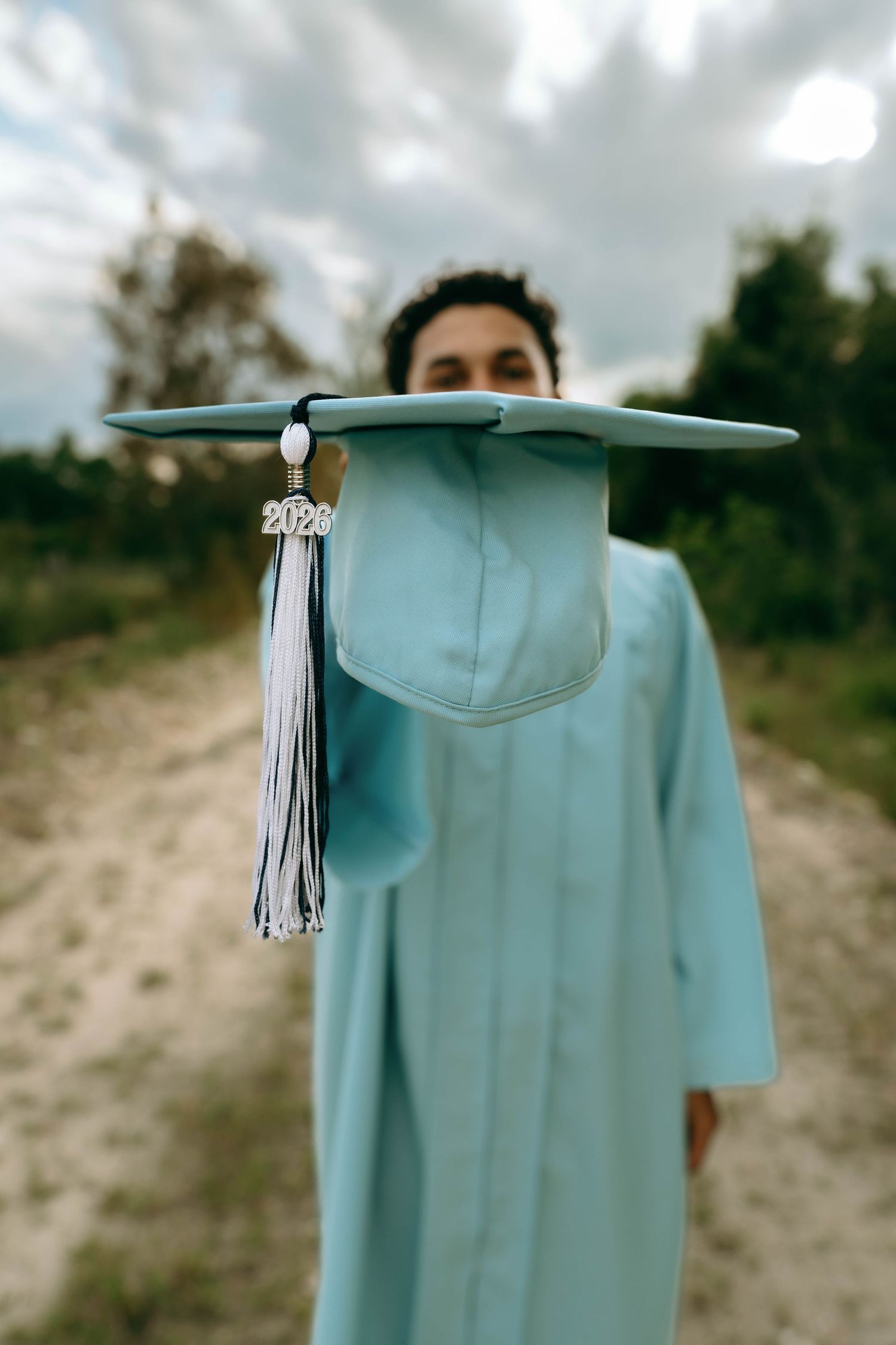 A graduate in a light blue cap and gown holds their graduation cap toward the camera on a sandy outdoor path. Senior Photos at Colt Farms