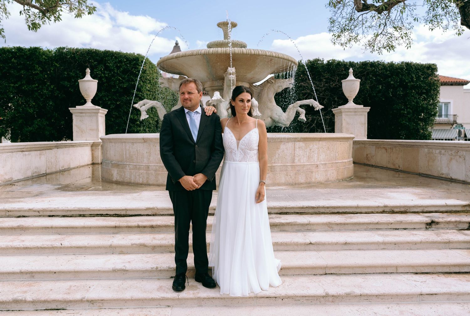 Bride and groom posing together in front of a grand decorative fountain on their wedding day.