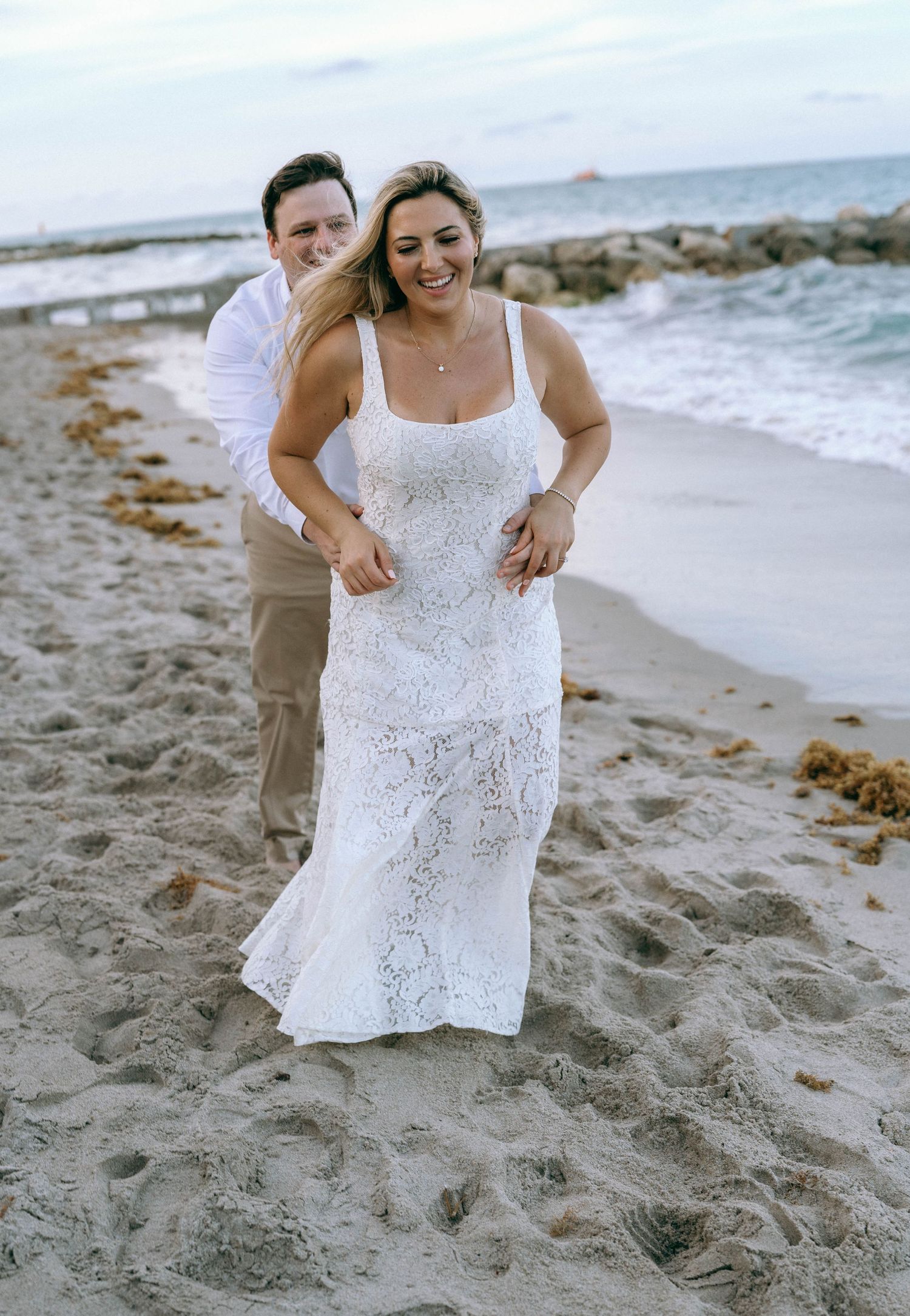 Happy couple playfully walking on sandy beach, woman wearing white lace wedding dress near ocean waves.