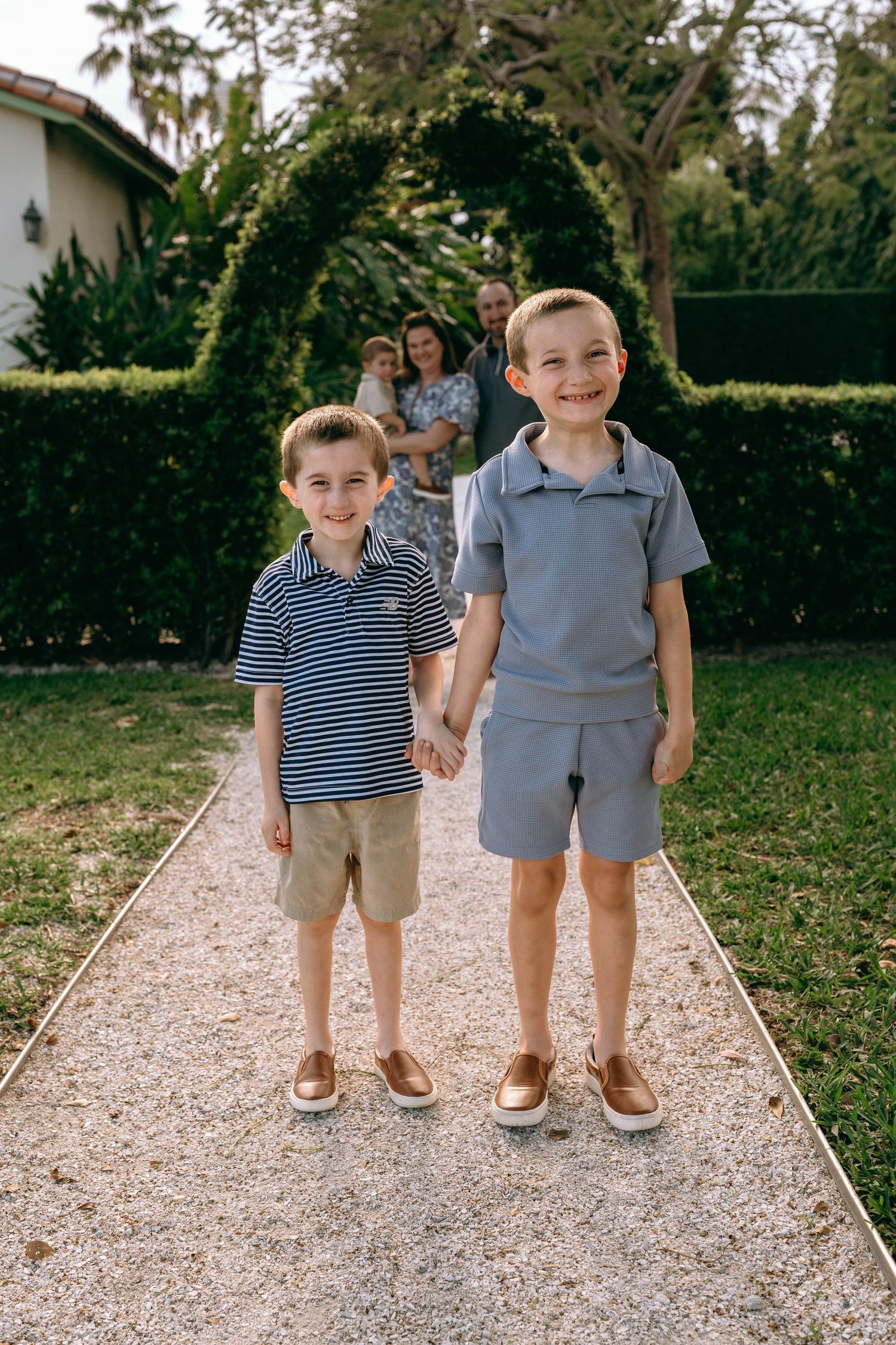 Two young brothers holding hands on garden path, smiling at camera with parents standing behind them.  Family photos at Bradley Park