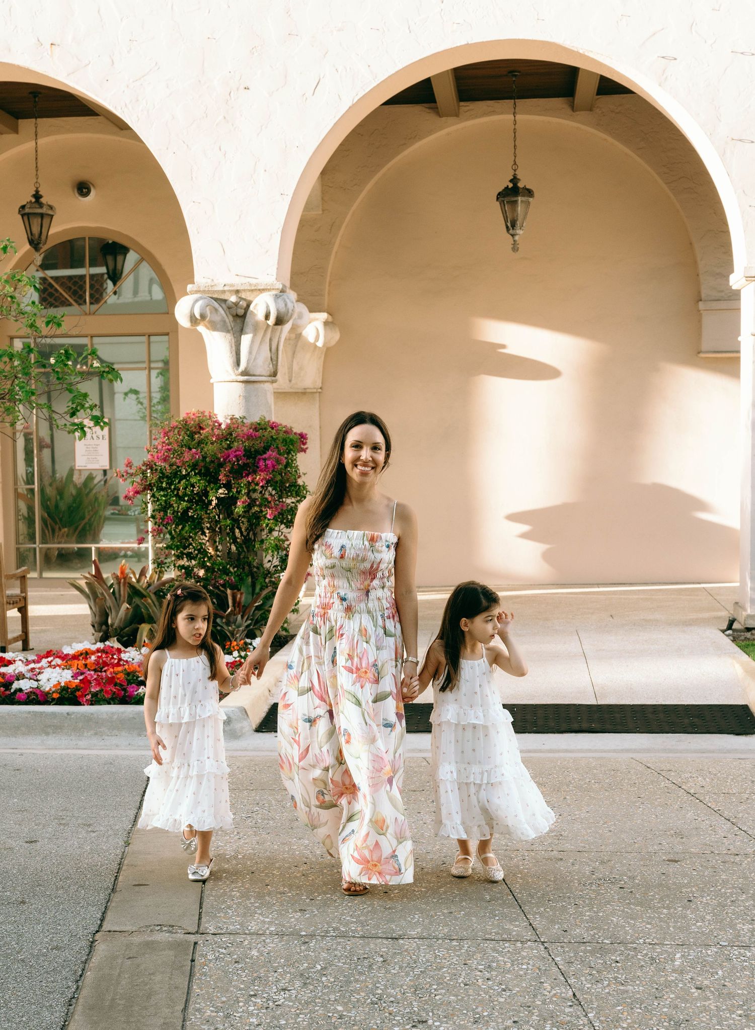 A smiling mother in a floral dress walks hand-in-hand with two girls in white dresses along a sunny colonnade. Worth Avenue, Palm Beach, FL