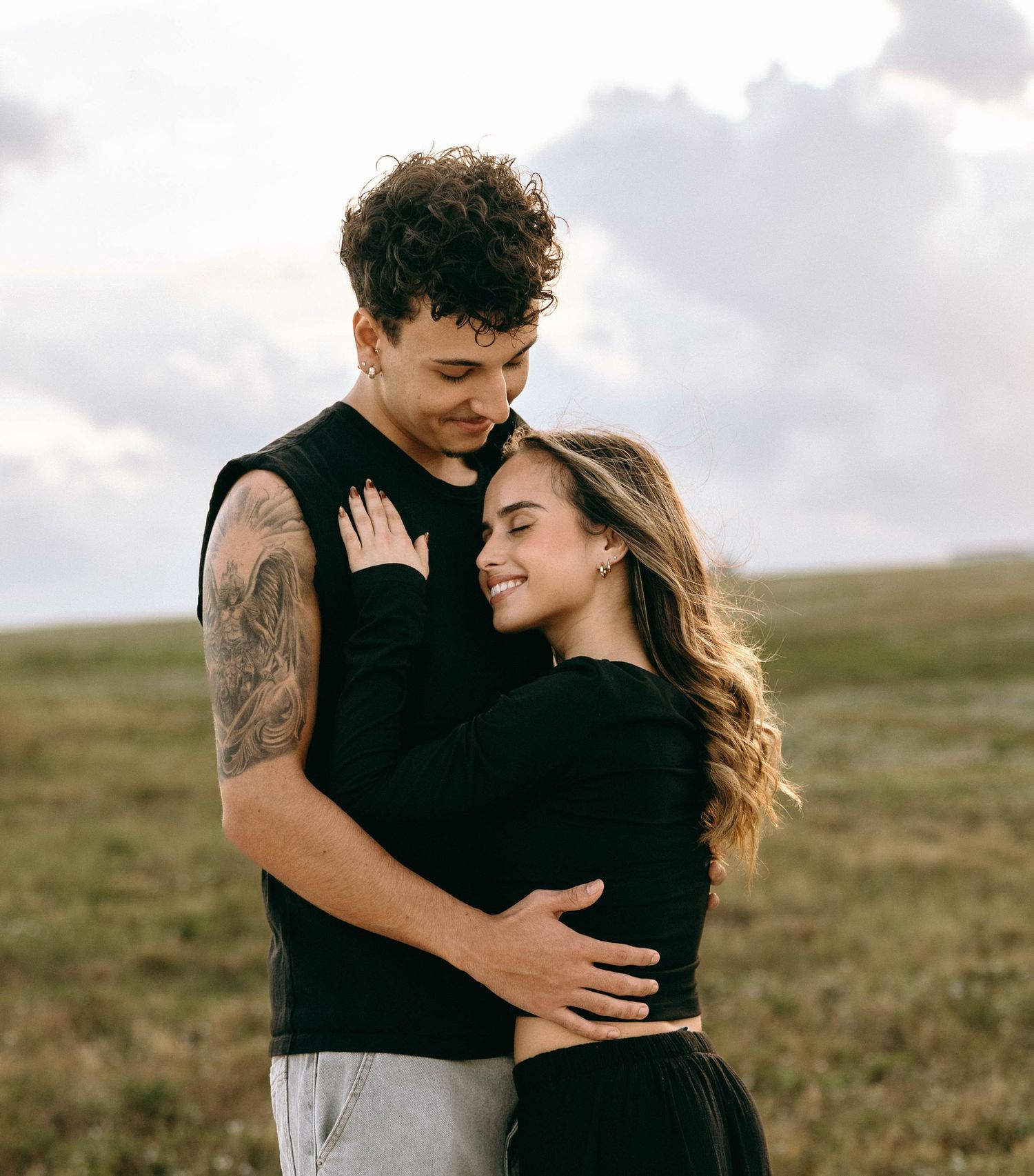Two people in black and gray clothing embrace tenderly in a grassy field.