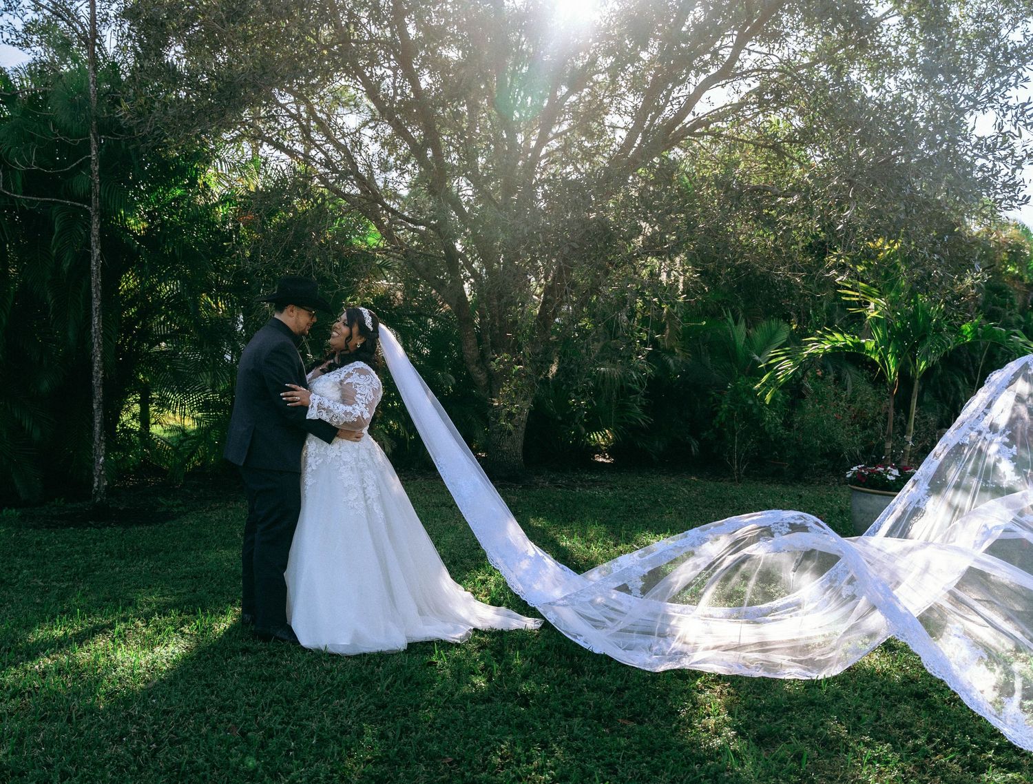 A flowing bridal veil creates dramatic silhouettes during an outdoor wedding photoshoot among trees.