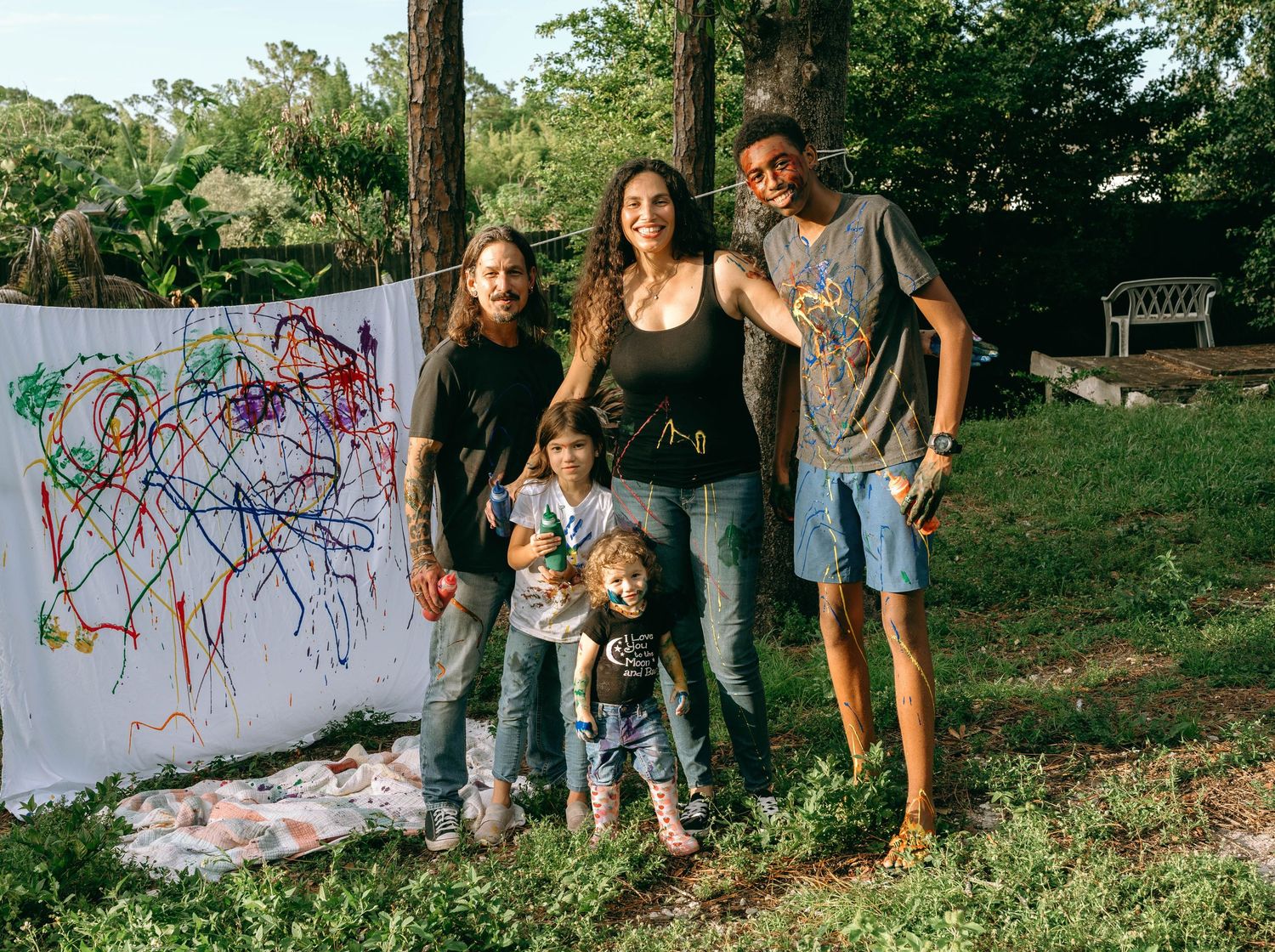 Family of five posing outdoors beside a large canvas with colorful artwork in a lush green backyard.