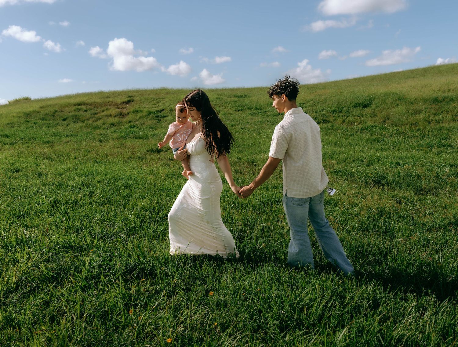 A young family holds hands walking through a lush green hillside meadow under a blue sky with clouds.