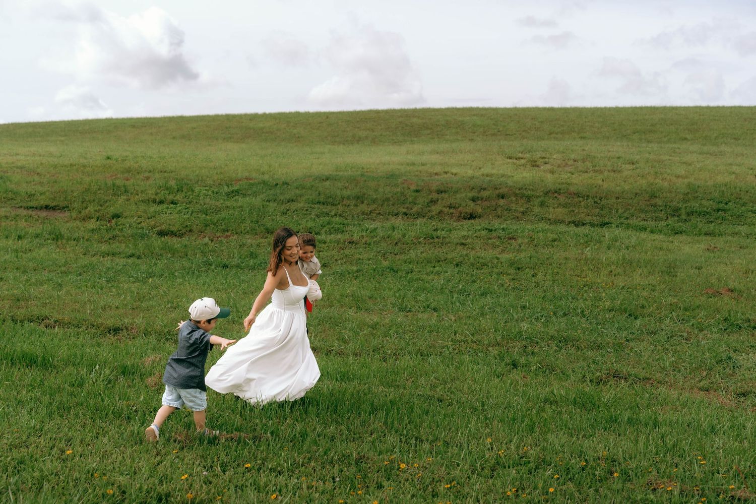A mother in a white dress holds hands with her toddler while playing in a lush green field under a cloudy sky.