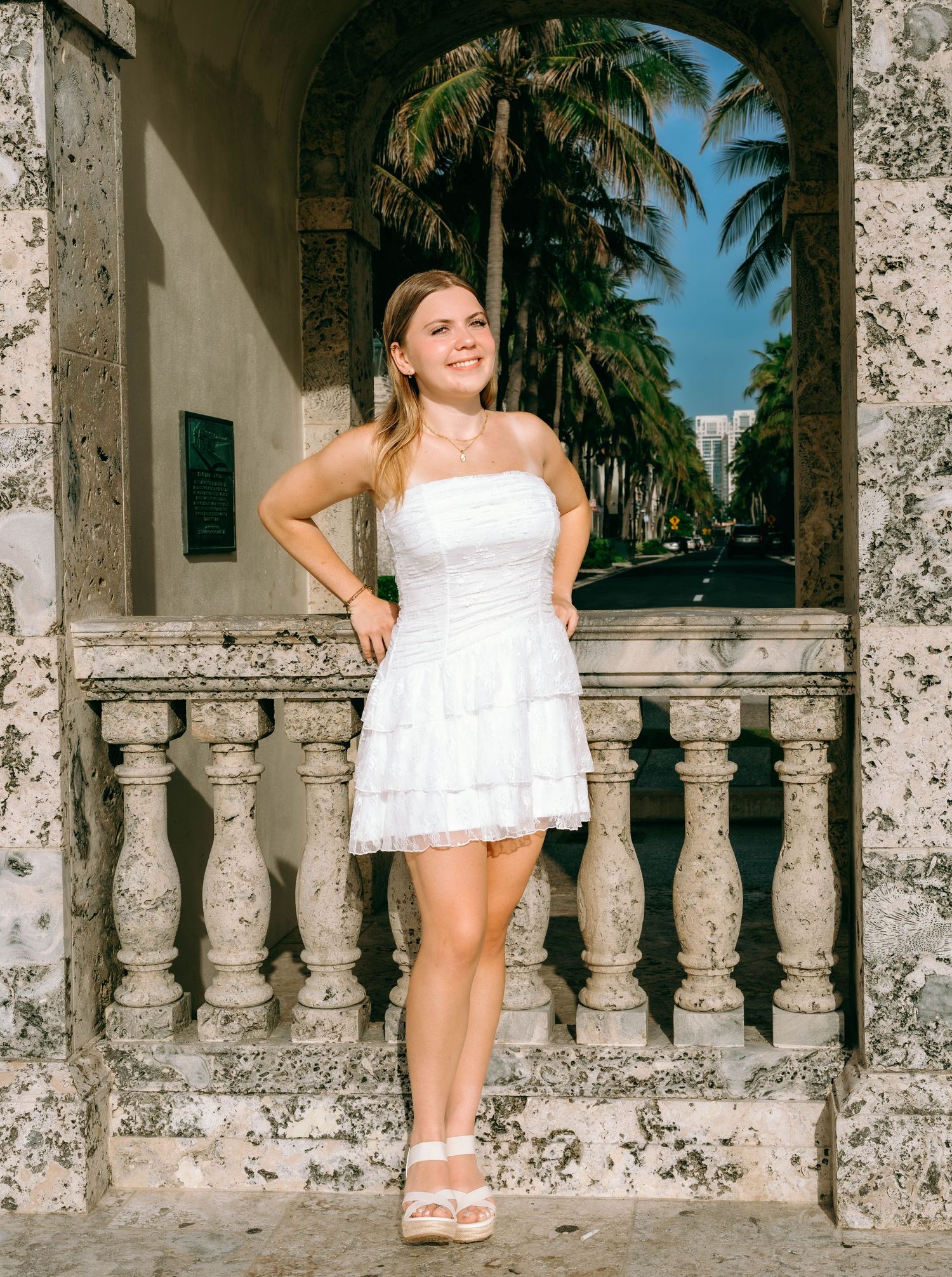 Young woman in white strapless mini dress posing at ornate stone balustrade with palm trees in background.