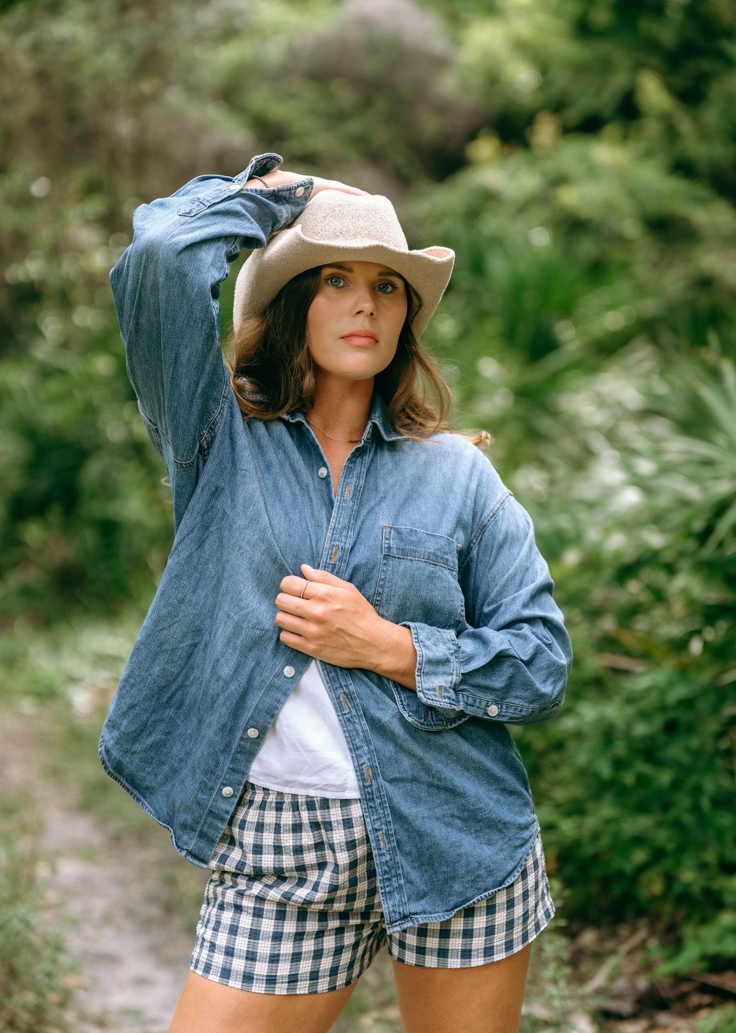 Woman in denim shirt, gingham shorts, and tan cowboy hat posing on a nature trail.