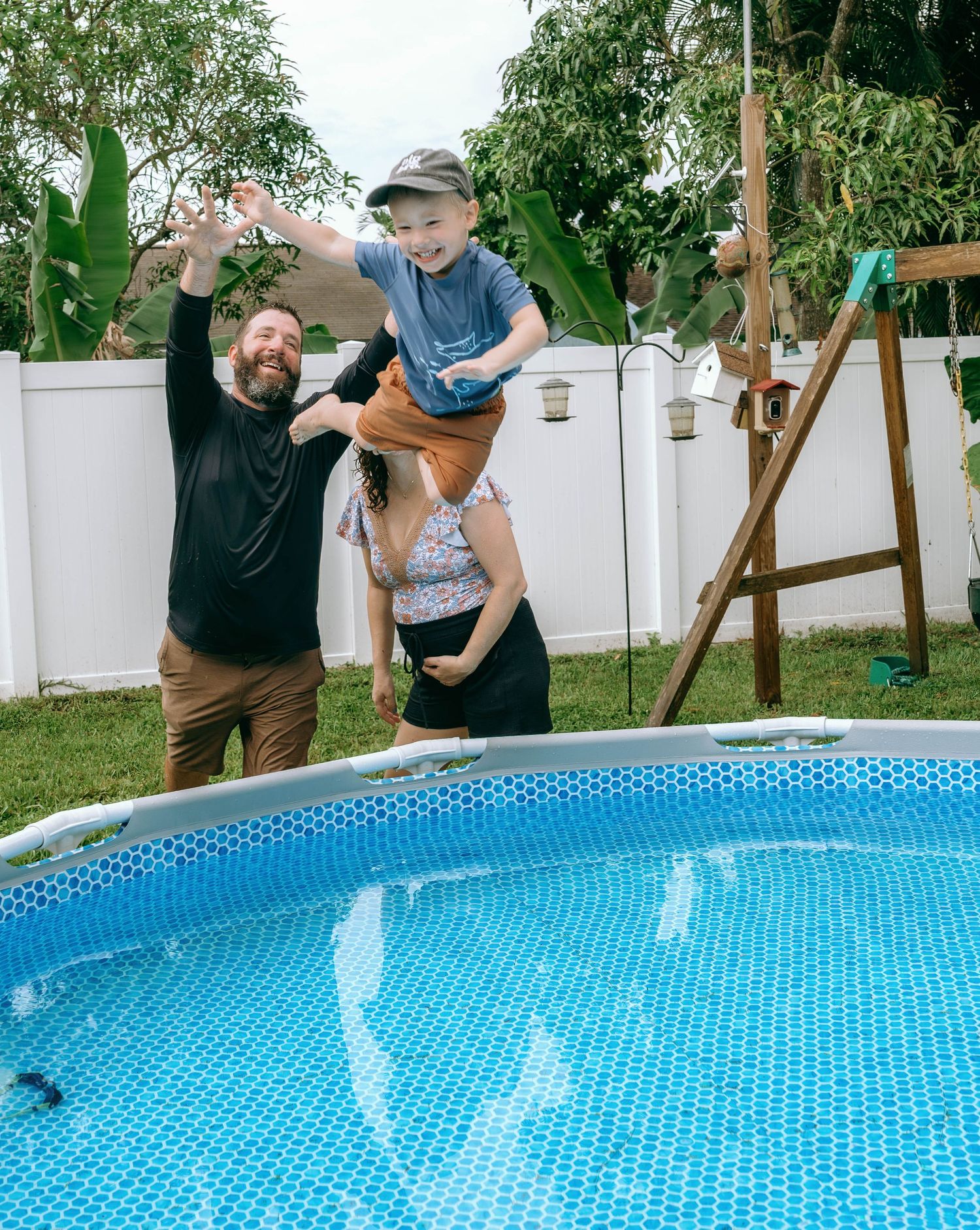 Family playing near an above-ground pool in backyard, lifting a child joyfully outdoors.
