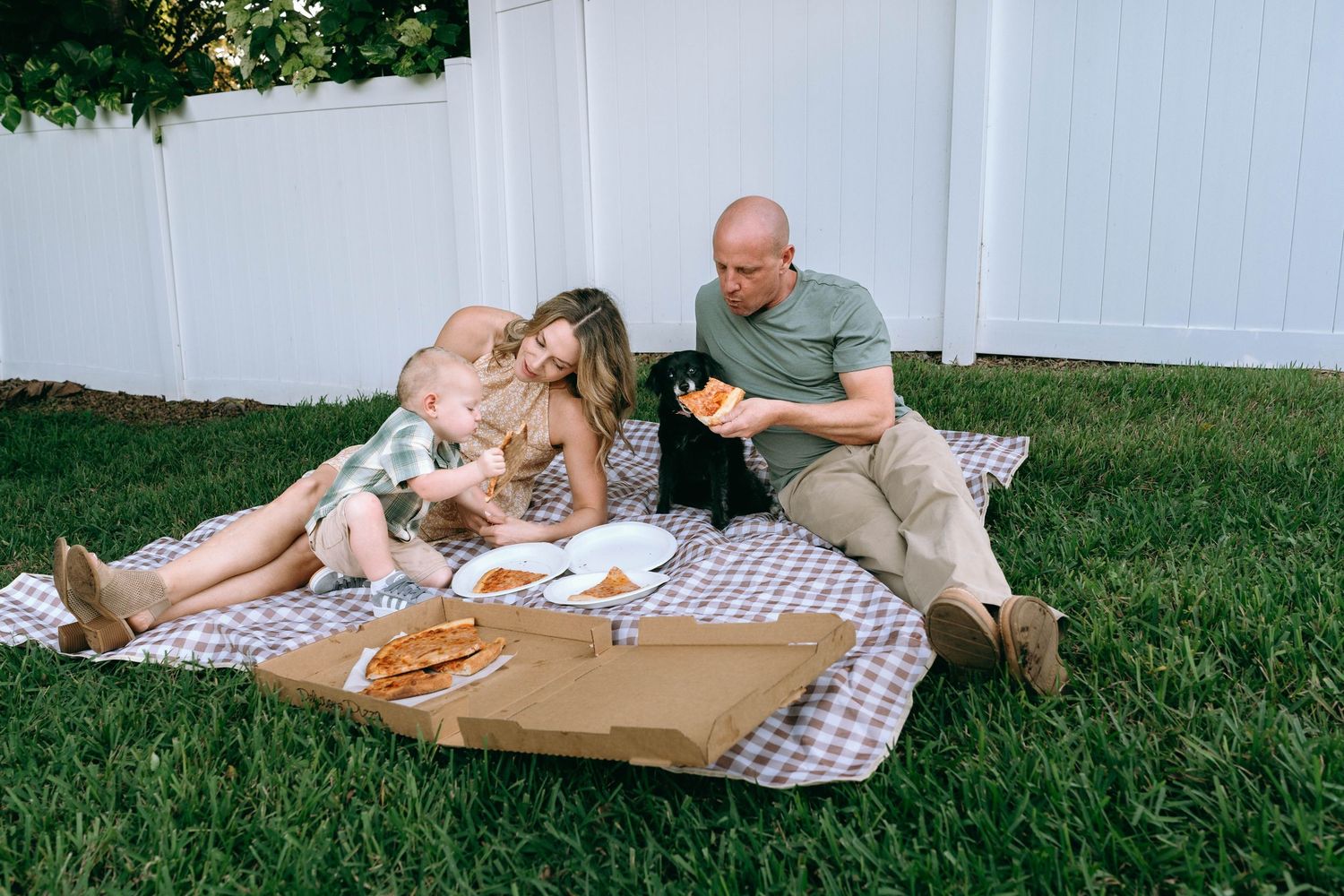 Family enjoying a casual outdoor picnic on a blanket by a white fence.