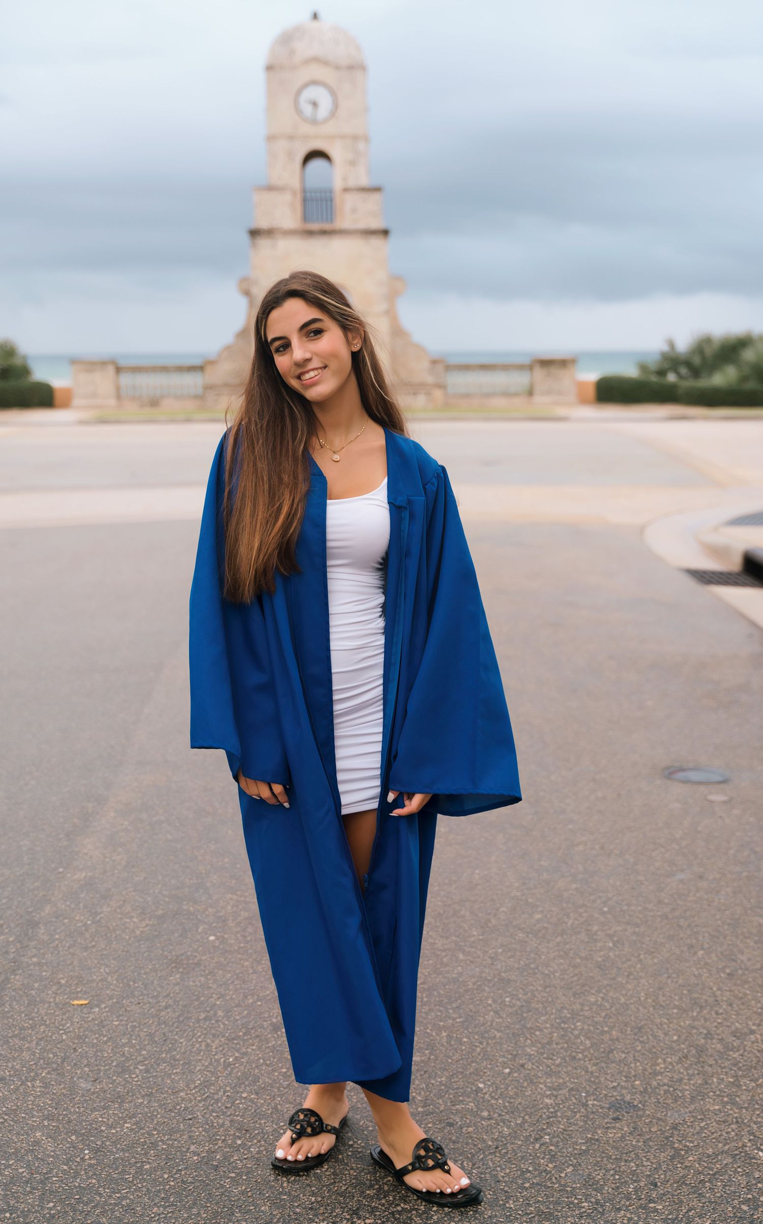 Graduate in blue academic regalia poses at memorial plaza under cloudy sky.
