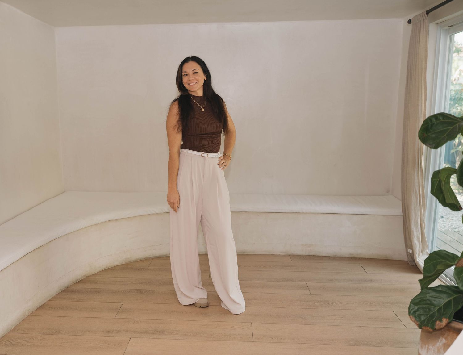 Woman in brown sleeveless top and wide-leg cream pants standing in a minimalist white room with wood floors.