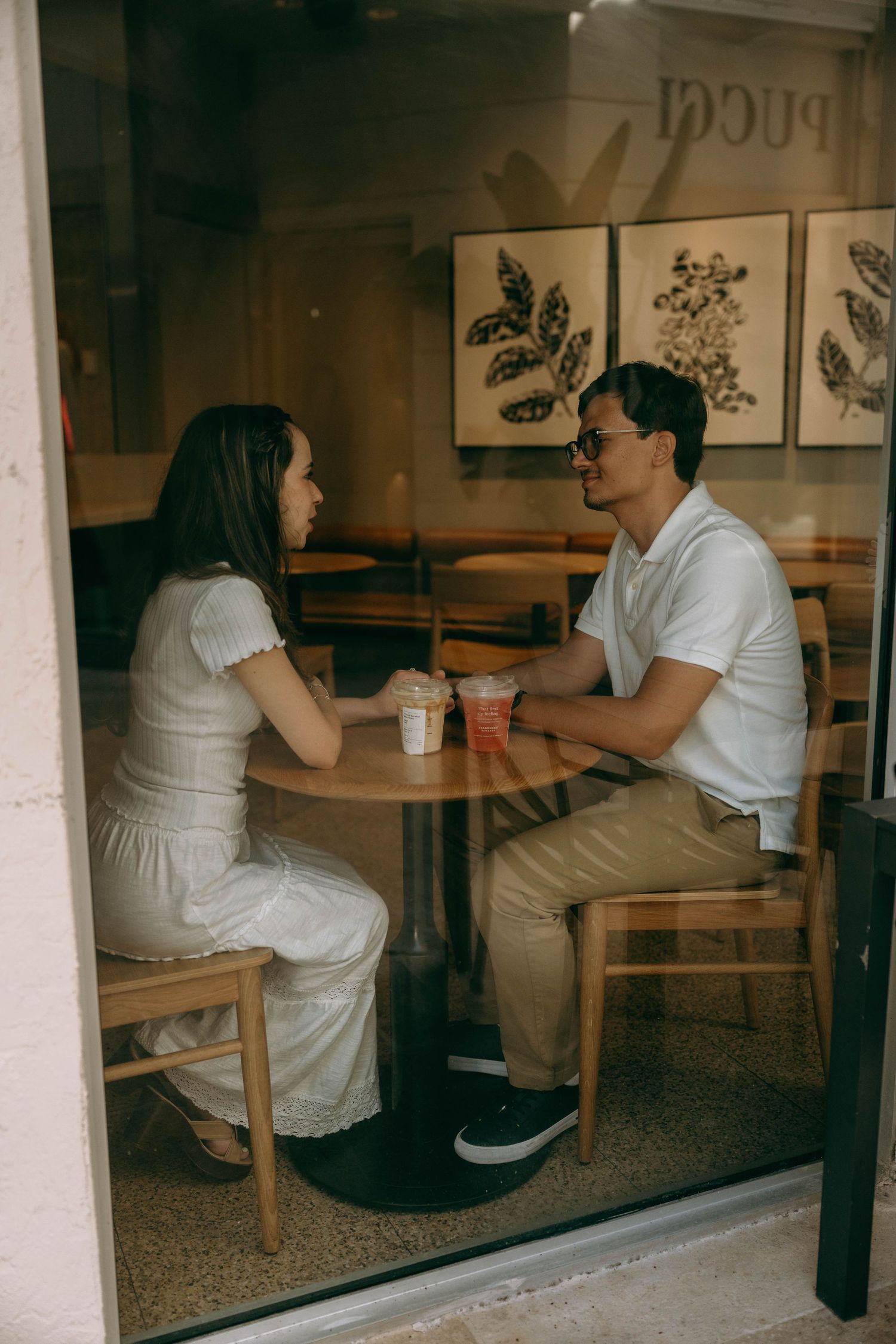 Couple on a coffee date sitting across from each other at a wooden table inside a cafe with iced drinks. Engagement photos on Worth Avenue