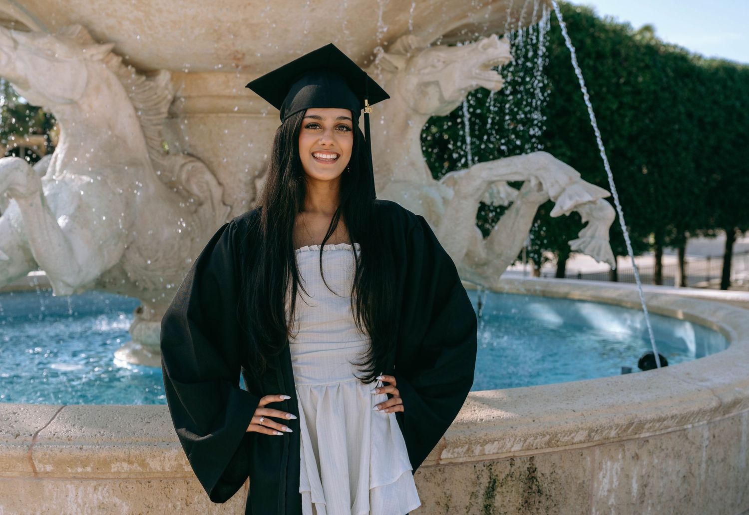 Smiling female graduate in black cap and gown poses proudly in front of a decorative fountain on Worth Avenue Palm beach.