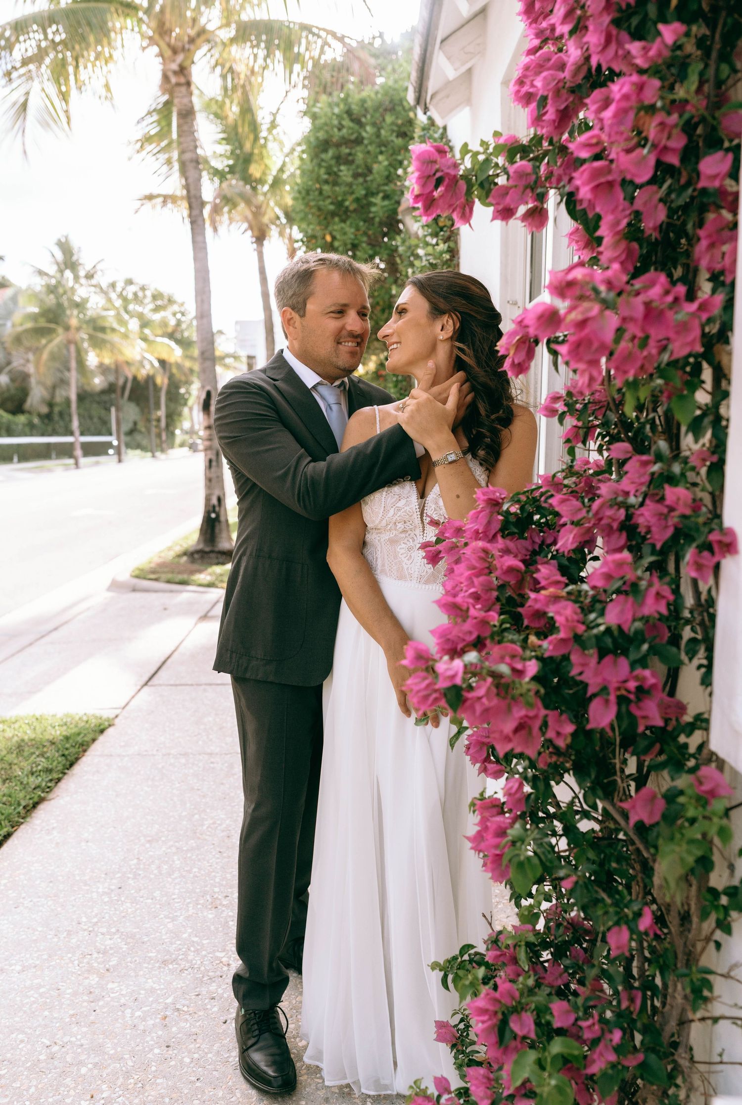Wedding couple embraces beside vibrant pink bougainvillea flowers on a sunny tropical sidewalk.