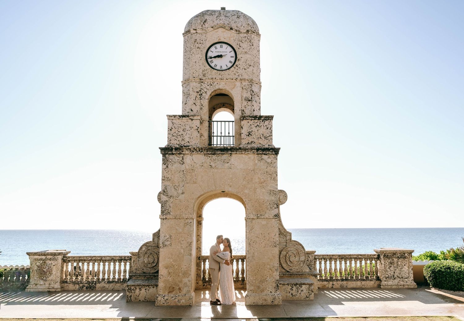 Couple kissing under historic clock tower arch with ocean view on their wedding day.