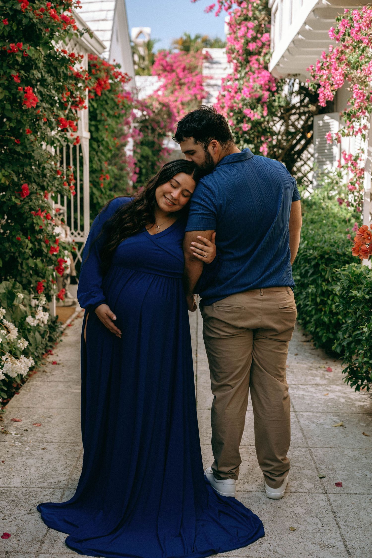 Pregnant woman in blue gown embraced by partner in floral garden during maternity photoshoot. Worth Avenue, Palm Beach, FL