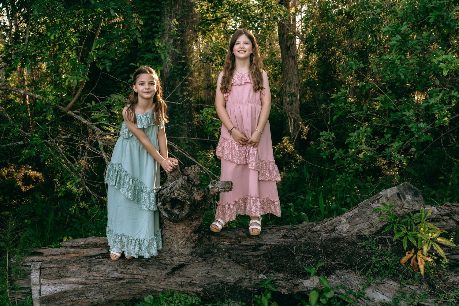 Two young girls in boho dresses standing on a fallen log in a sunlit forest setting. Colt Farms