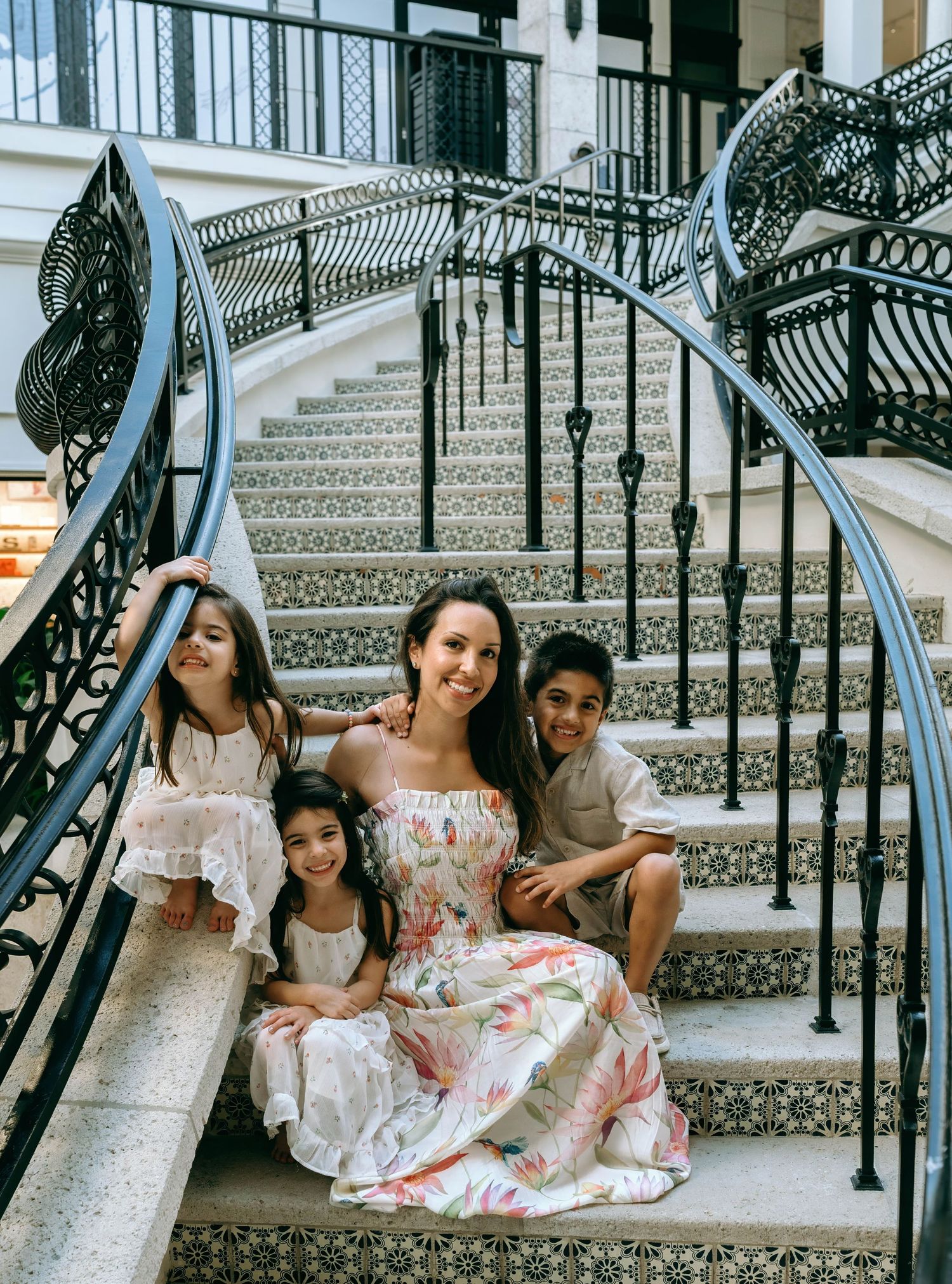 A happy mother in a floral maxi dress poses with her three children on elegant tiled staircase with iron railings.