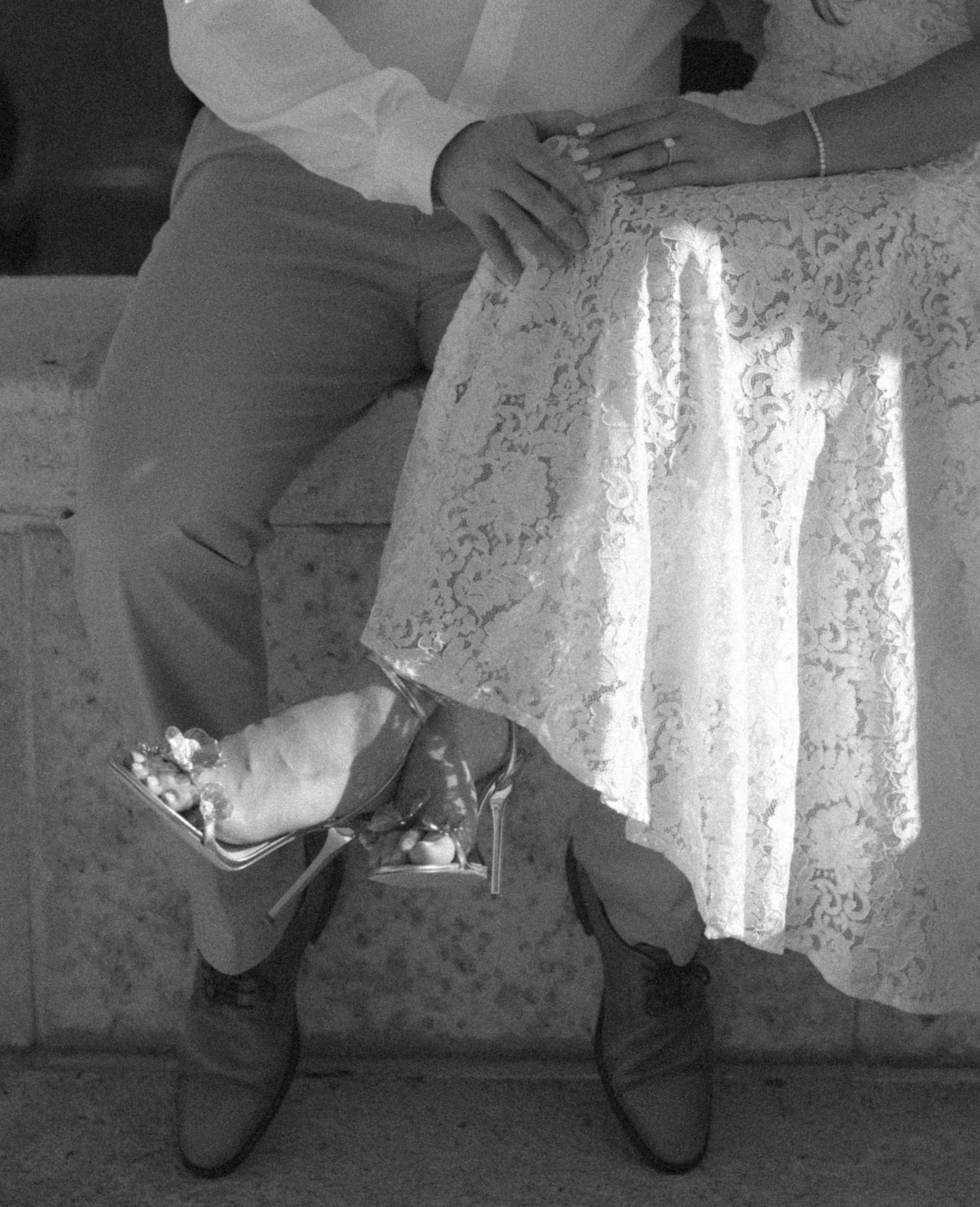 Black and white photo of a couple sitting together, woman in lace dress and heels, man in dress pants and boots.