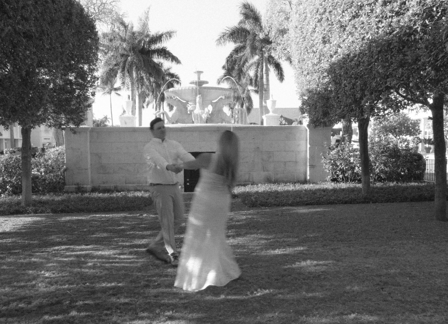 Black and white photo of a couple dancing outdoors near a fountain surrounded by palm trees.