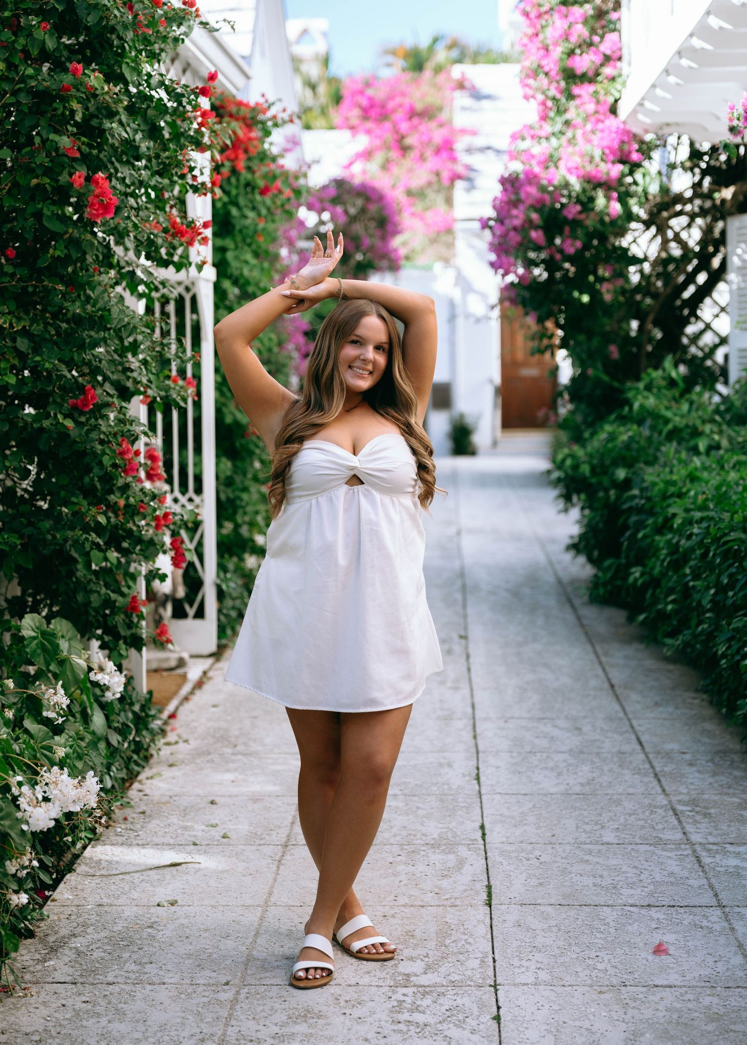 Woman in white strapless cutout dress posing in flower-lined walkway with pink and red bougainvillea.