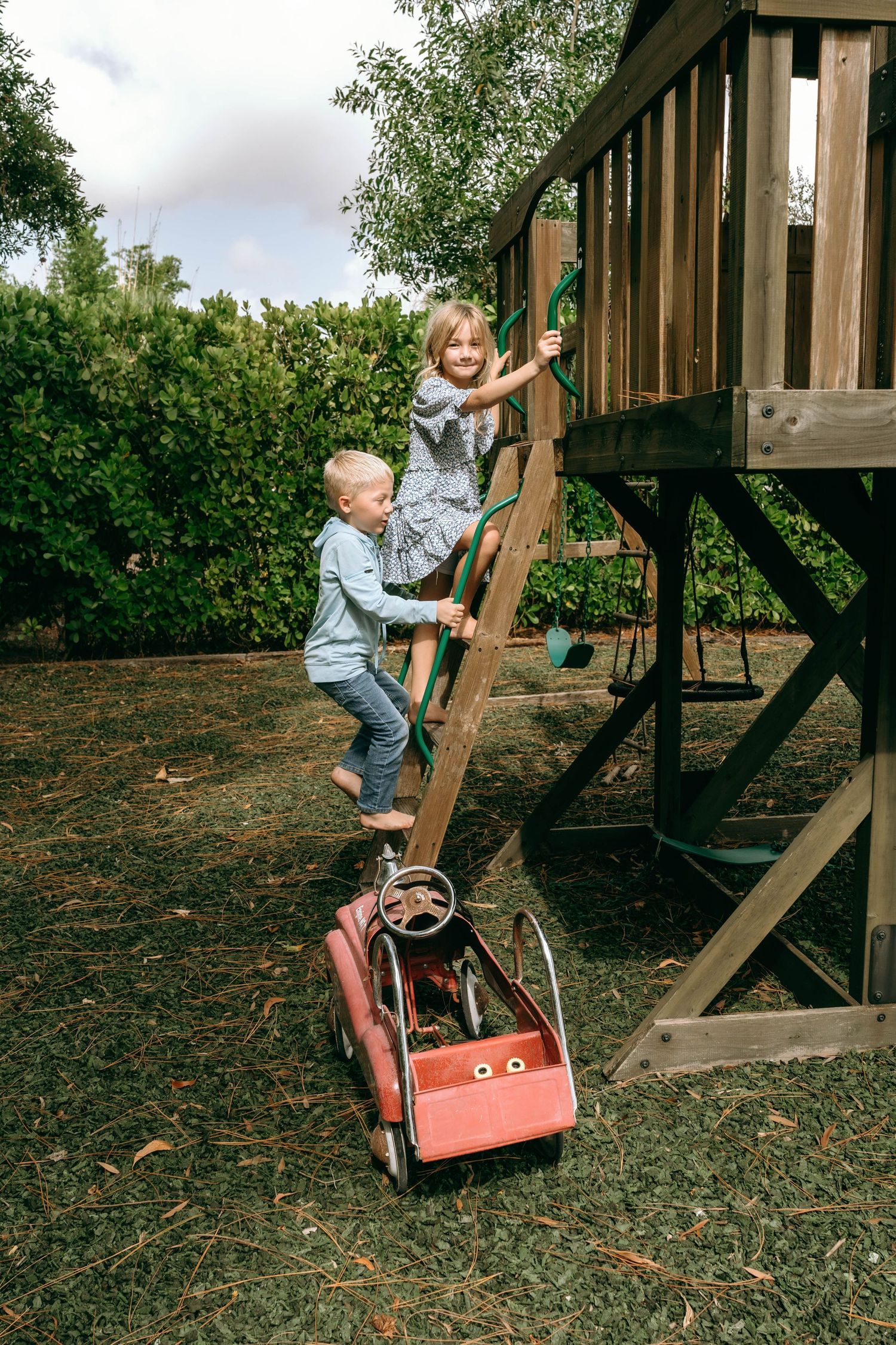 Two young children climbing wooden ladder to backyard treehouse playset with red wagon below.
