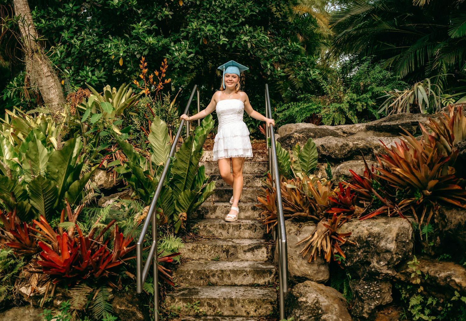 Woman in white dress and blue hat descends lush tropical garden stone stairs surrounded by colorful exotic plants.